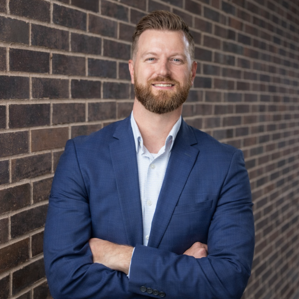 A man with a beard in a blue suit smiling and crossing his arms against a gray background.