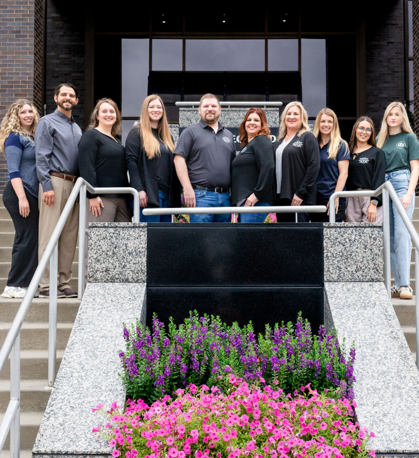 Group of nine people standing on steps outside a building, smiling for the photo. There are stairs and colorful flowers in the foreground.