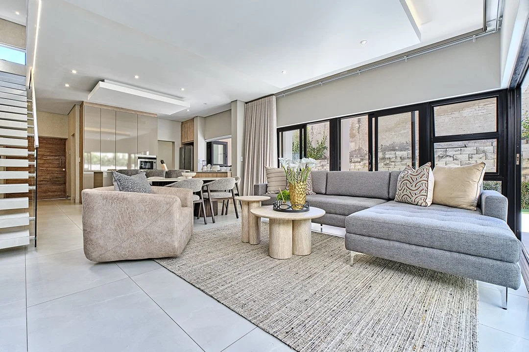Modern living room with gray sectional sofa, beige armchair, wooden coffee tables, and a large window with black frames, bright natural light, neutral decor, and a textured area rug.