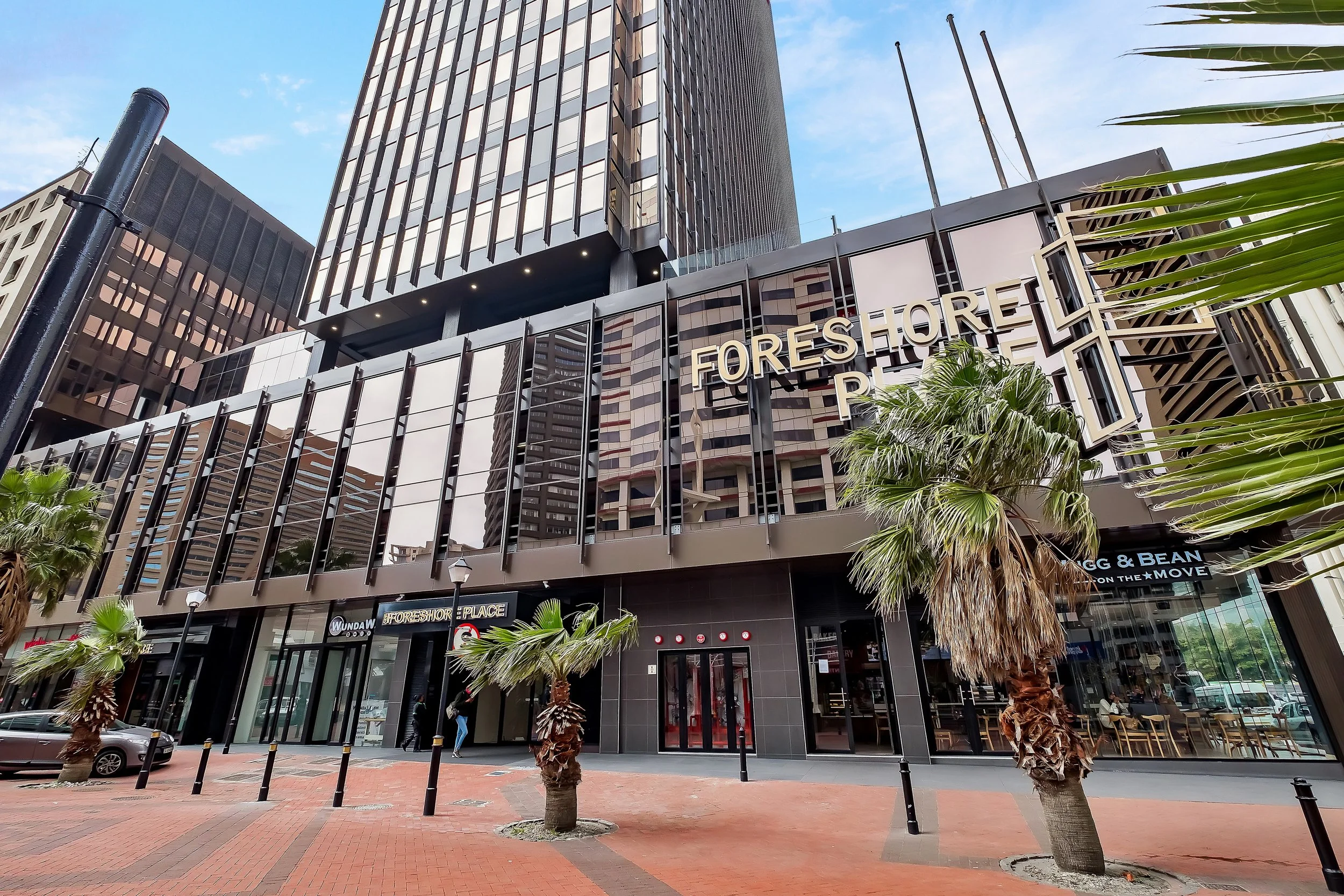 A modern multi-story commercial building with glass and dark panel exterior, displaying the sign 'Foreshore Place'. There are palm trees, a sidewalk, and people walking nearby, with a clear blue sky above.