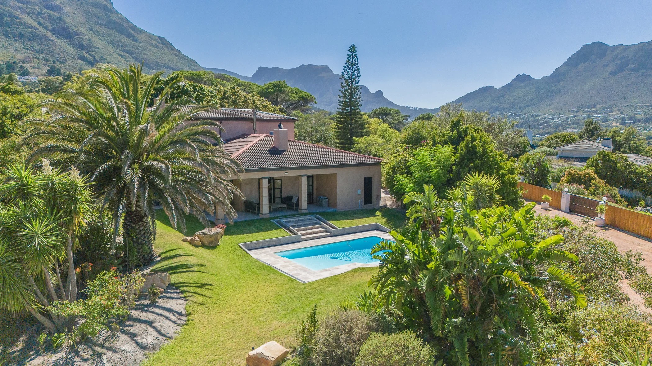 Monterey Cape town , A backyard with a swimming pool, surrounded by lush green plants, trees, and landscaped grass, with a house that has a covered patio, and mountains in the background under a clear blue sky.