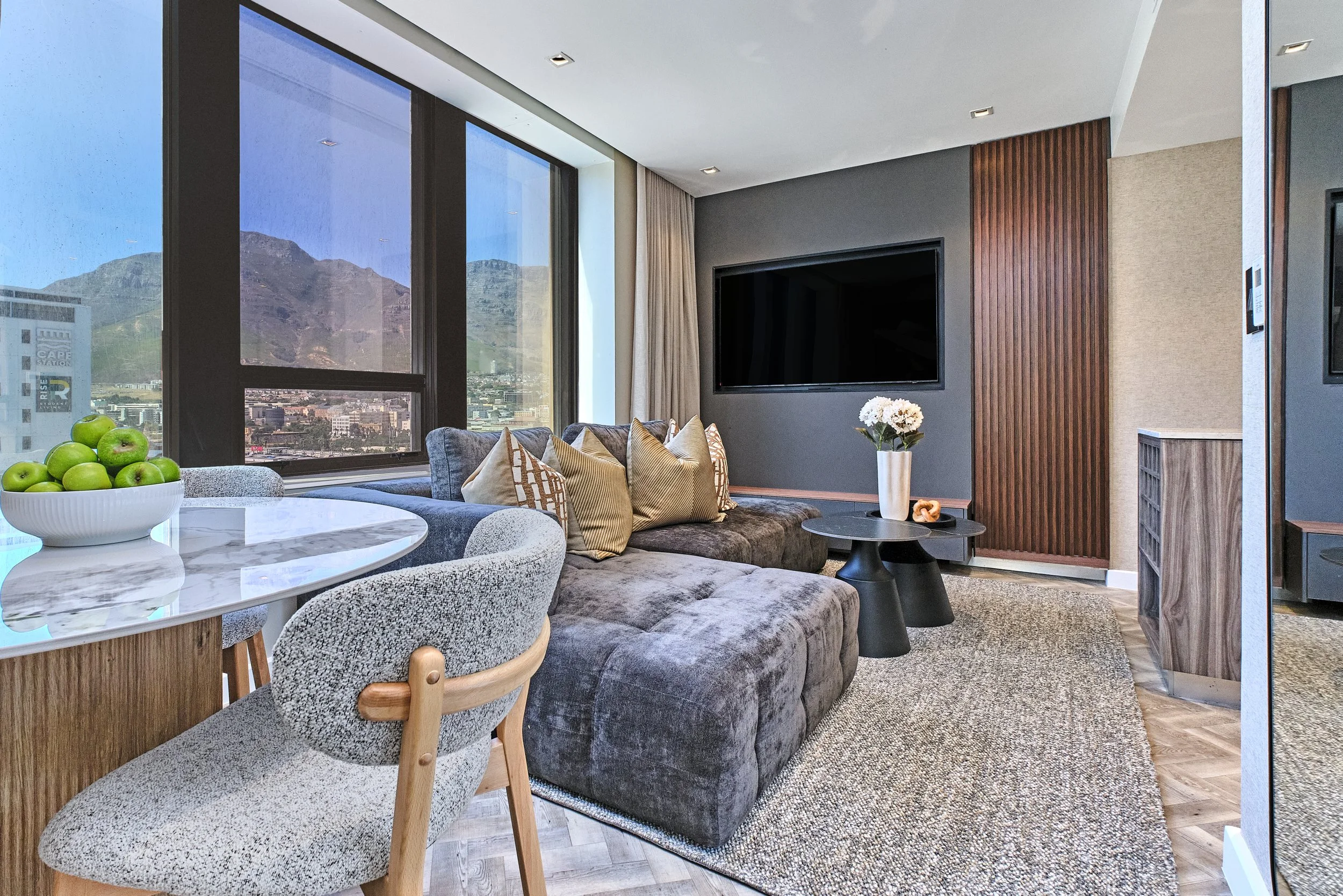 Modern living room with large windows showing mountain views, a gray sofa with multiple cushions, a coffee table with a white flower vase, a tv mounted on the wall, and a bowl of green apples on a dining table.