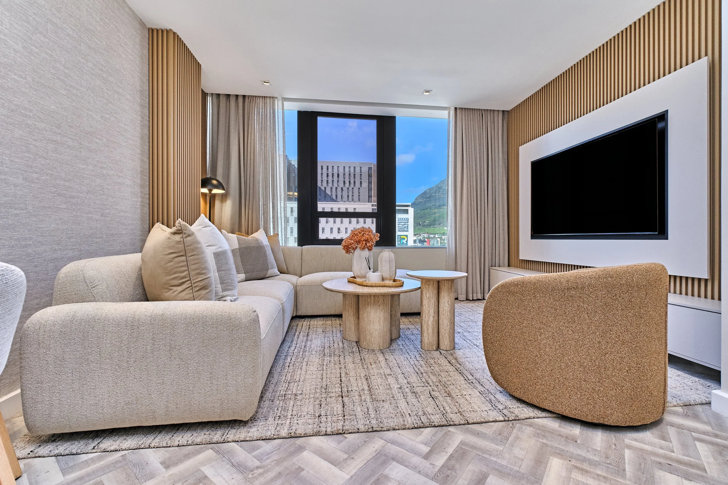 A modern living room with a beige curved sofa, a round wooden coffee table, a textured beige and gray area rug, a flat-screen TV on a wall with vertical wooden slats, large windows with beige curtains, mountain views, and decorative vases with dried flowers.