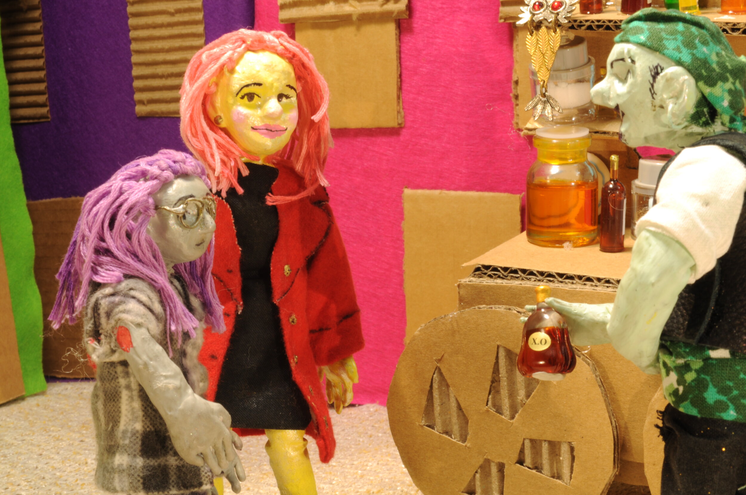  A claymation scene of a mother and daughter speaking to a potion master. Mother with pink hair and a red coat is standing to the left with her daughter with purple hair and glasses next to her. On the right side is the potion master with a green ban