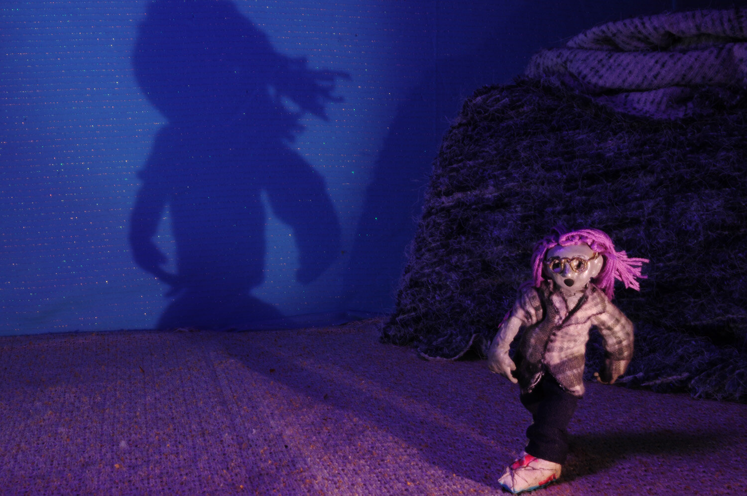  A claymation scene of a young girl with purple hair running. Behind her is a dark purple rock, and her shadow can be seen against the blue wall. 
