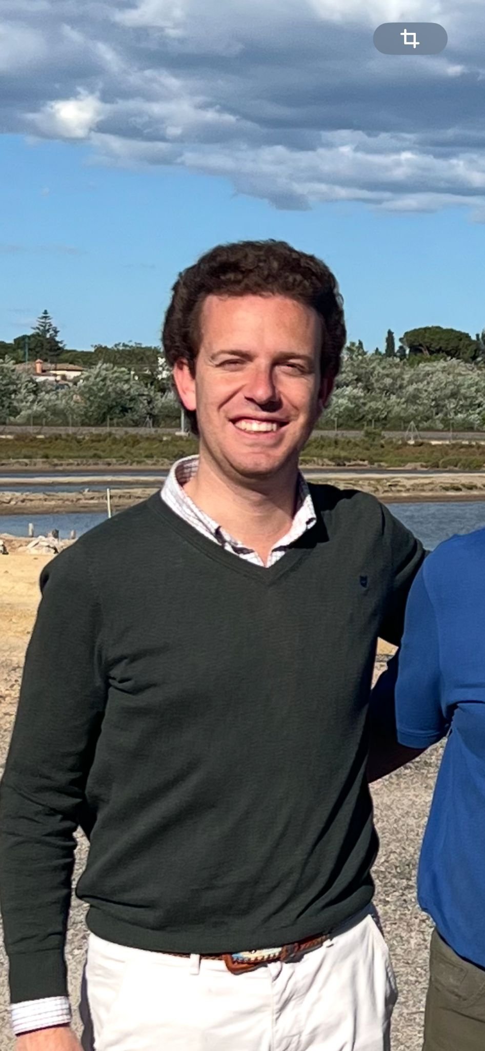 A smiling man with dark, curly hair wearing a green sweater over a checkered shirt, standing outdoors near water under cloudy skies.