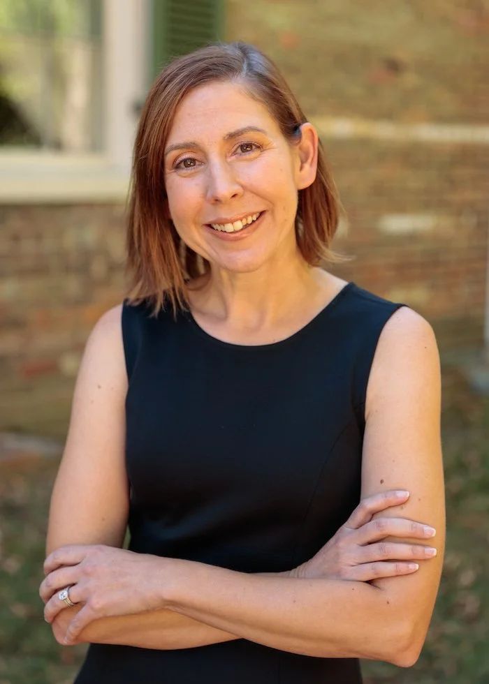 A woman with shoulder-length reddish-brown hair, wearing a sleeveless black dress, standing outdoors with her arms crossed and smiling at the camera.