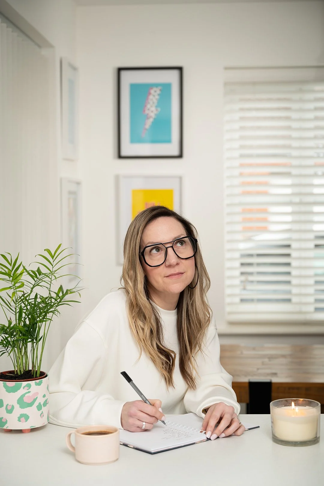 A woman with long blonde hair, glasses, and a white sweater sitting at a desk, writing in a notebook. There is a potted plant, a cup of coffee, and a lit candle on the desk, with framed artwork on the wall behind her.