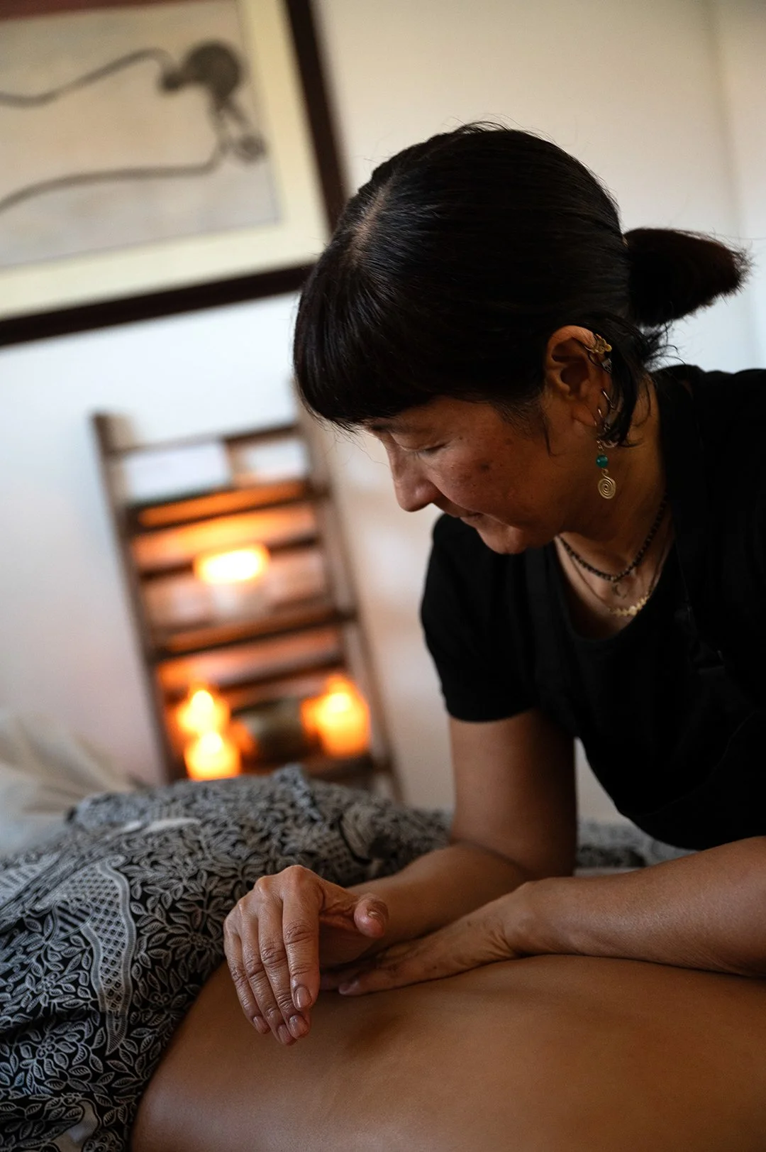 A woman is giving a massage or healing therapy to a person lying face down on a bed in a dimly lit room with candles and framed artwork on the wall.