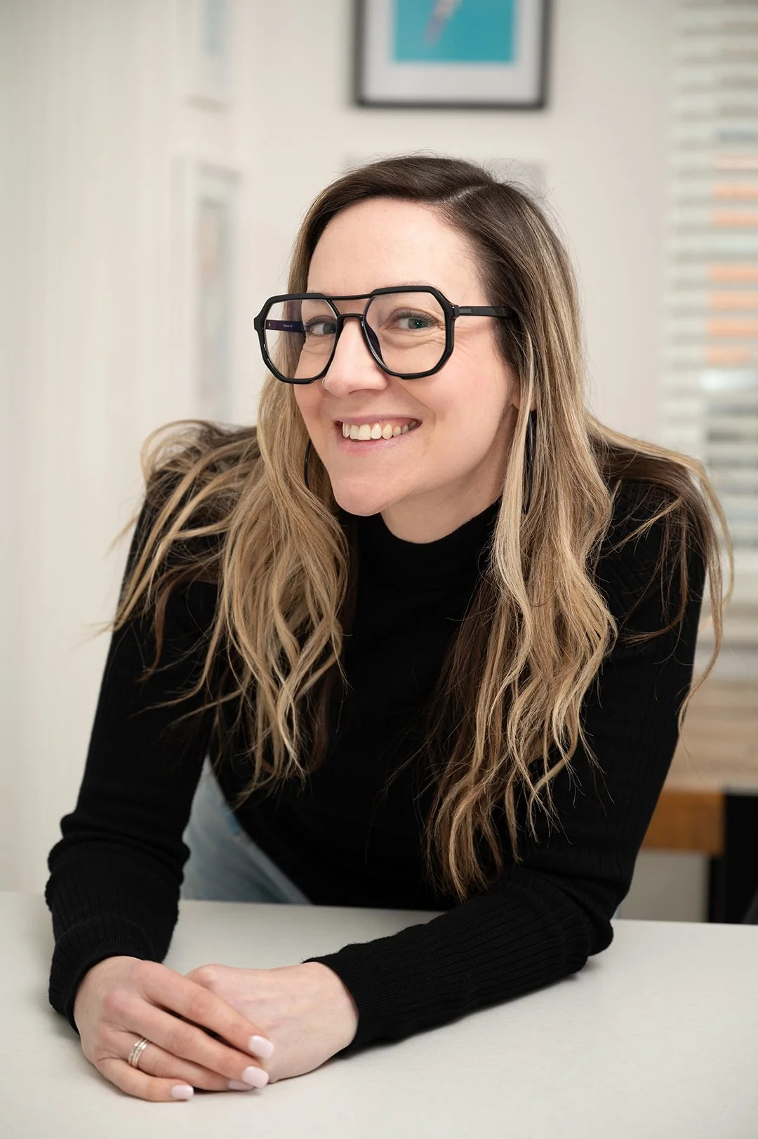 A woman with long wavy hair wearing large black glasses and a black turtleneck, smiling and leaning forward with arms resting on a white table.