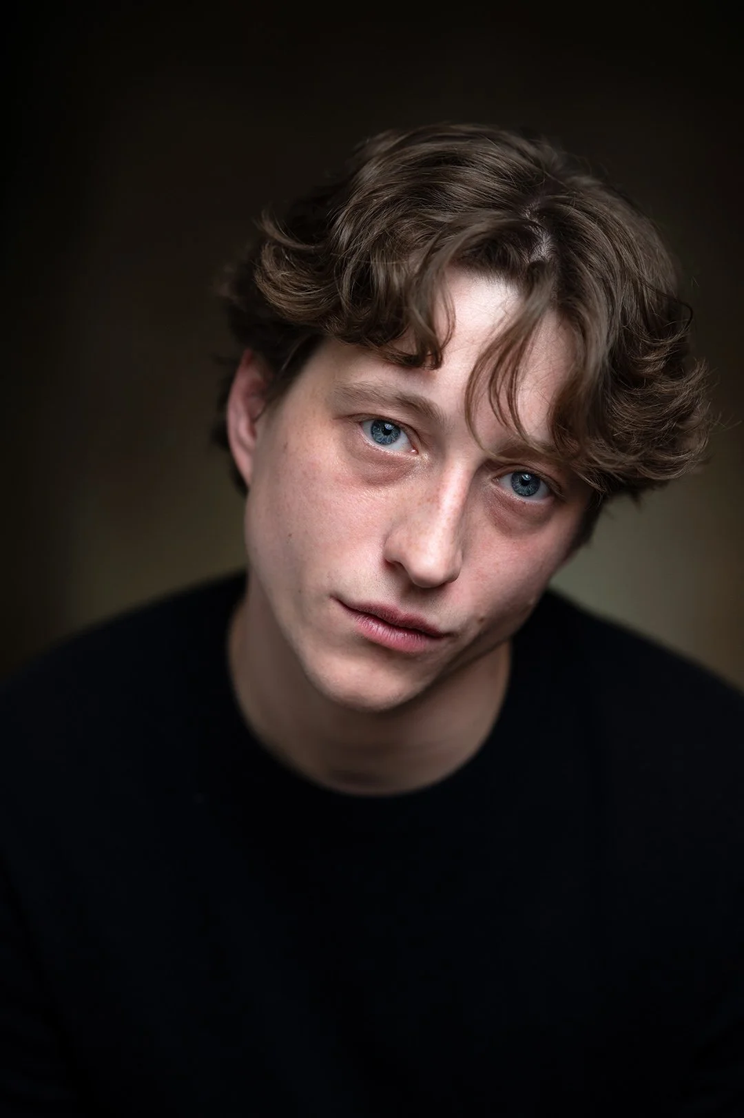 Portrait of a young man looking directly at the camera wearing a black jumper and against a dark brown background. 
