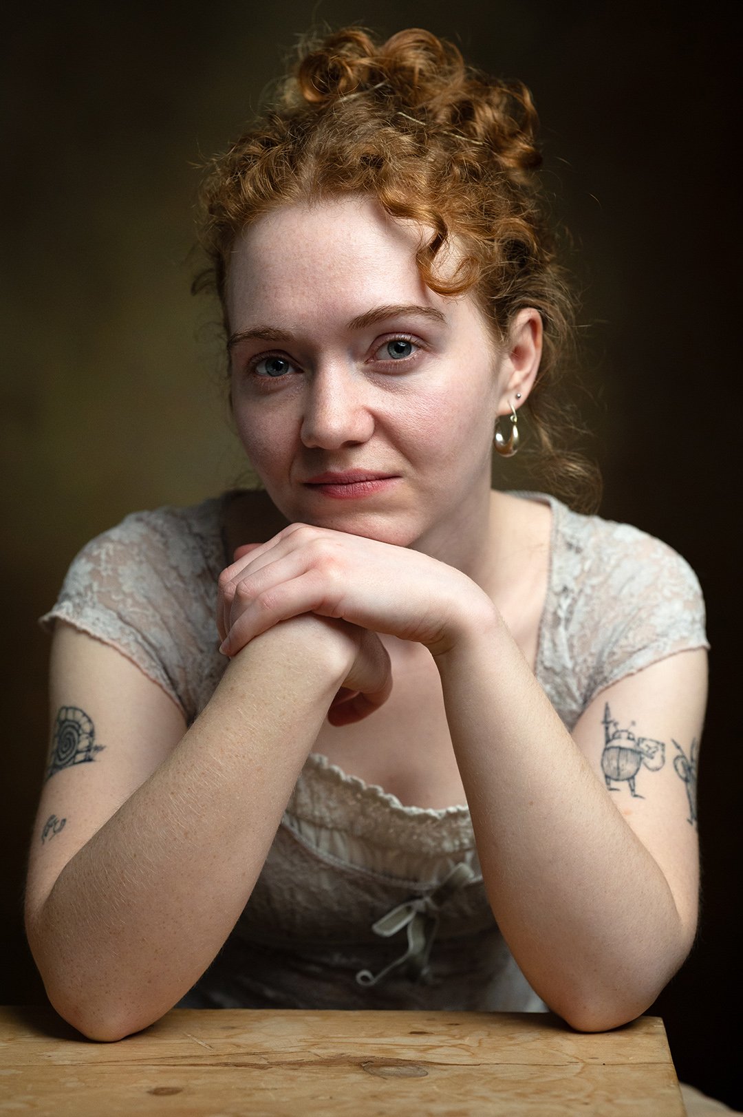 A woman with red curly hair and tattoos on her arms, wearing a lace top and earrings, resting her chin on her hands, looking at the camera in a dark studio setting.
