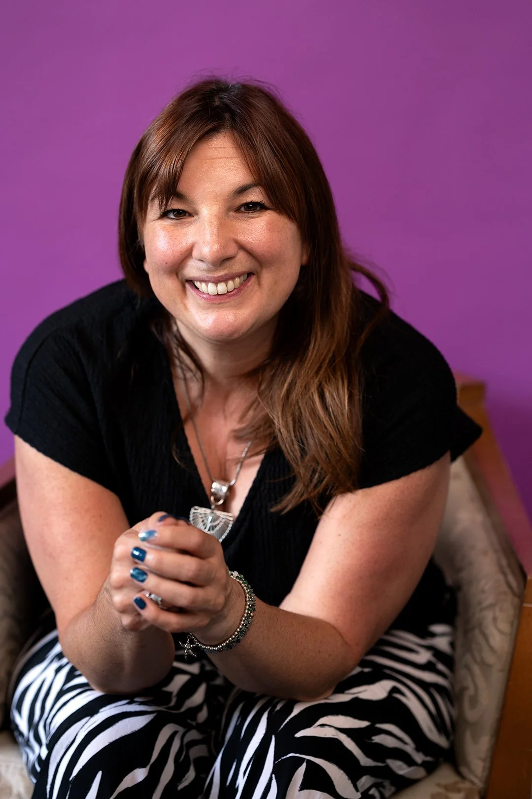 A female wearing a black top and stripey skirt photographed smiling sitting in a chair with a purple backdrop