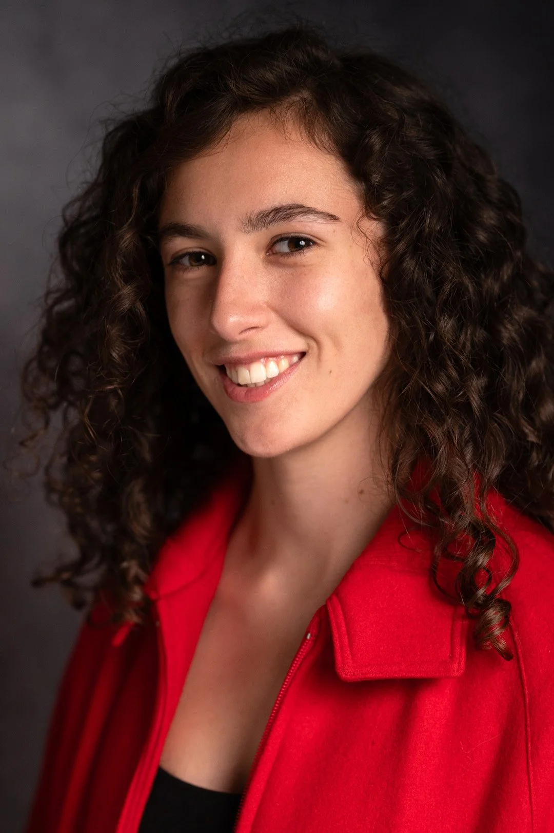 A young woman with dark curly hair and a red jacket, smiling, 