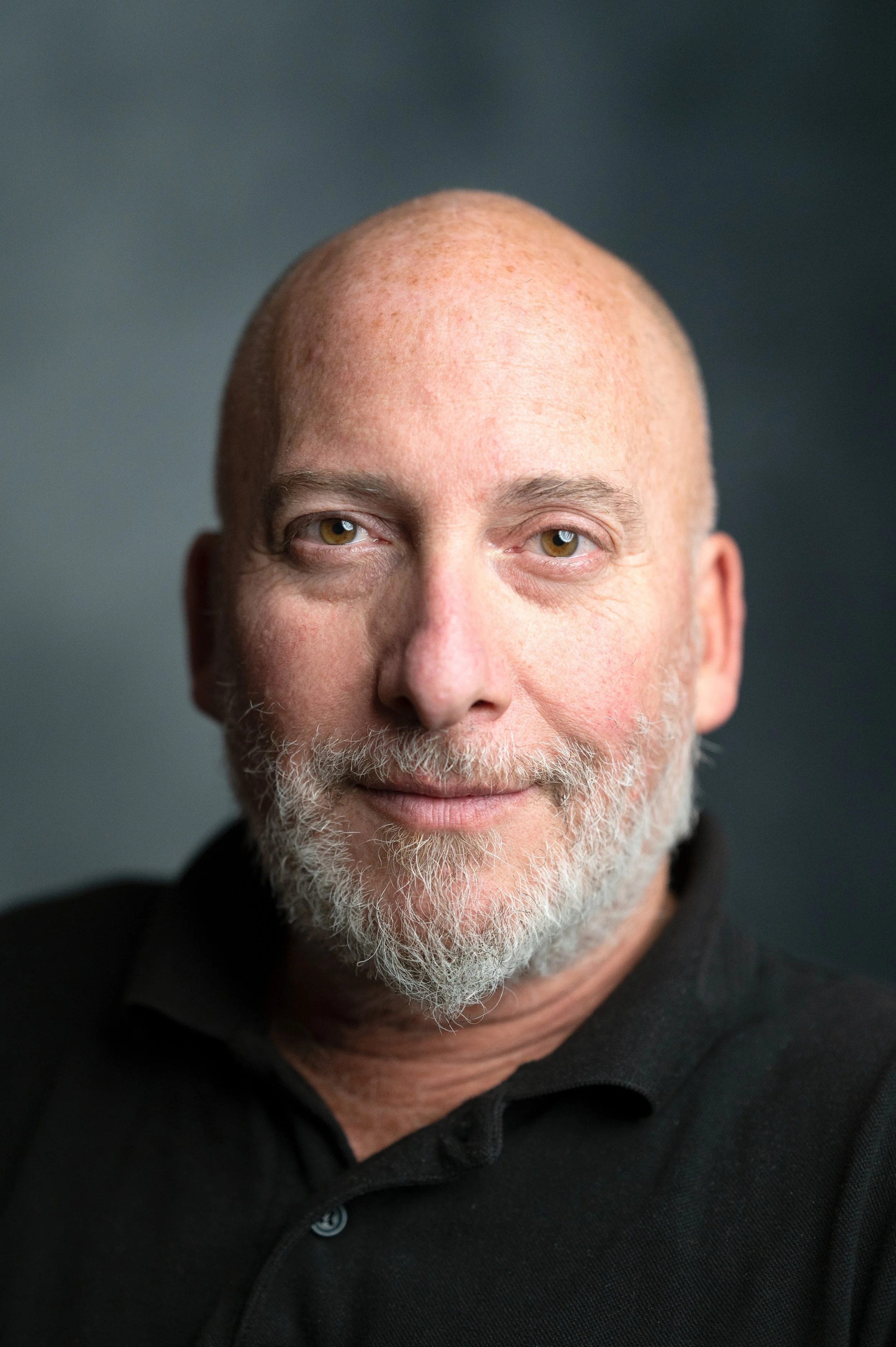 A male professional  with bald head wearing a black top with collar and a white beard. He is photographed against a grey backdrop looking straight at the camera with a slight smile. 