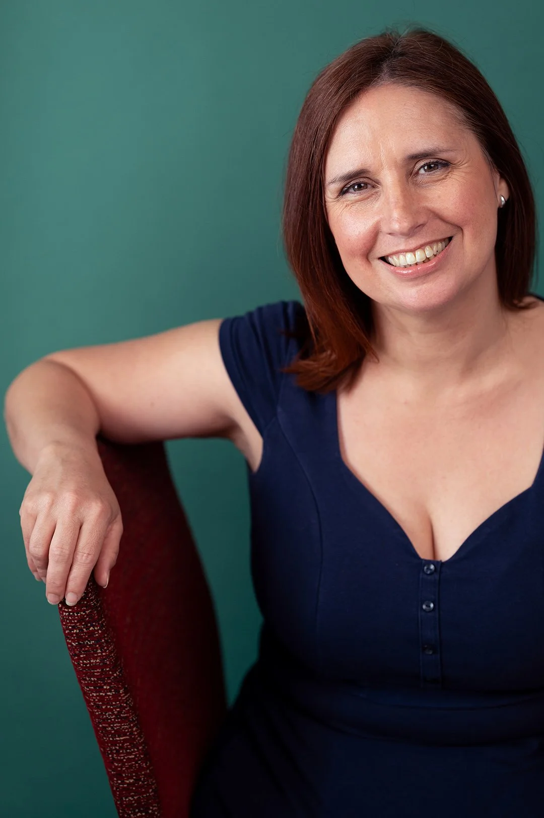 Female entrepreneur smiling straight at the camera wearing a blue dress and leaning one arm along the top of a chair