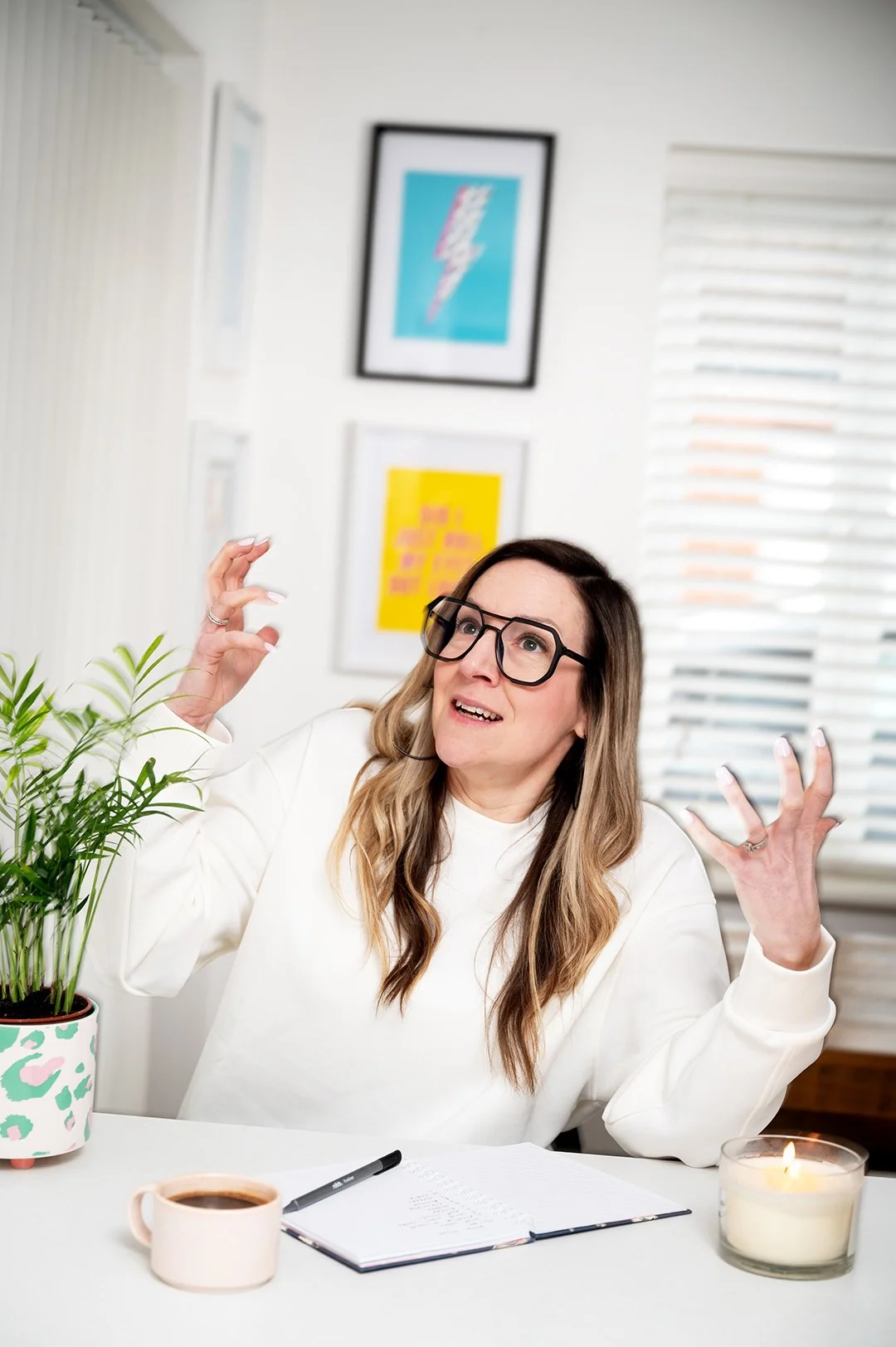 A woman with long brown hair wearing glasses and a white sweater sitting at a desk with a notebook, a pen, a cup of coffee, a lit candle, and a potted plant. She appears to be gesturing or explaining something with a surprised or expressive face, in 