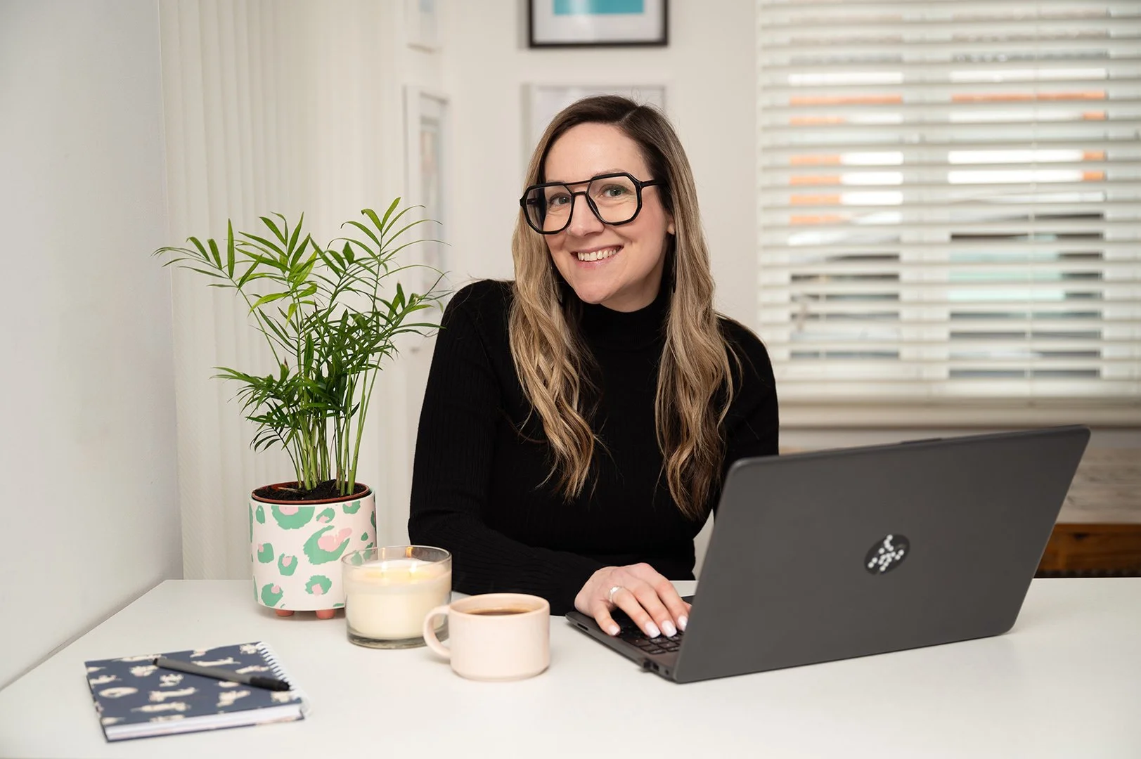 A woman with long, wavy hair and glasses sitting at a white desk, smiling at the camera, with a laptop, potted plant, candle, mug, and notebooks on the desk, in a bright room with window blinds.