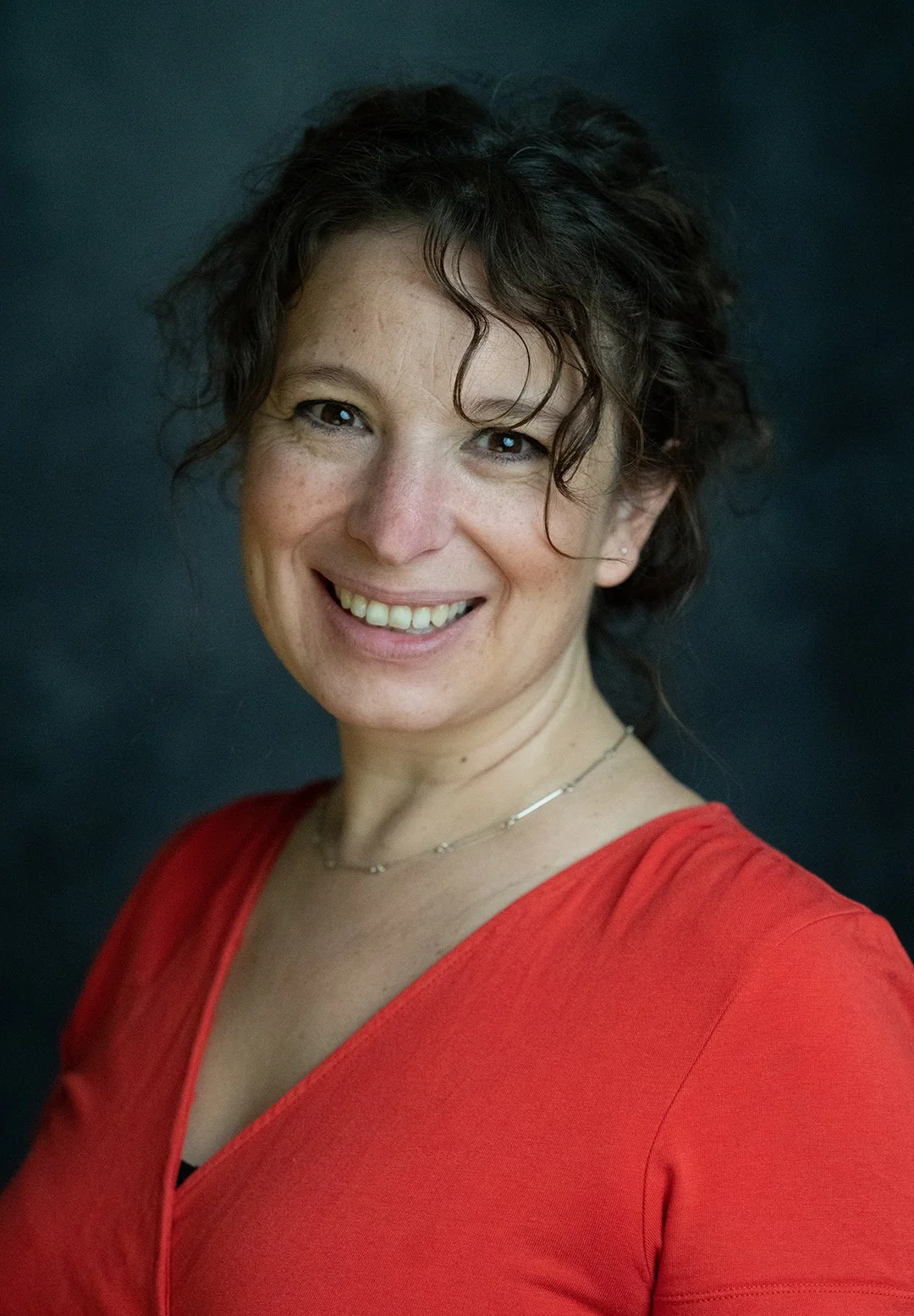 Woman wearing red top smiling at the camera with dark short hair.  Photographed against a dark grey backdrop. 