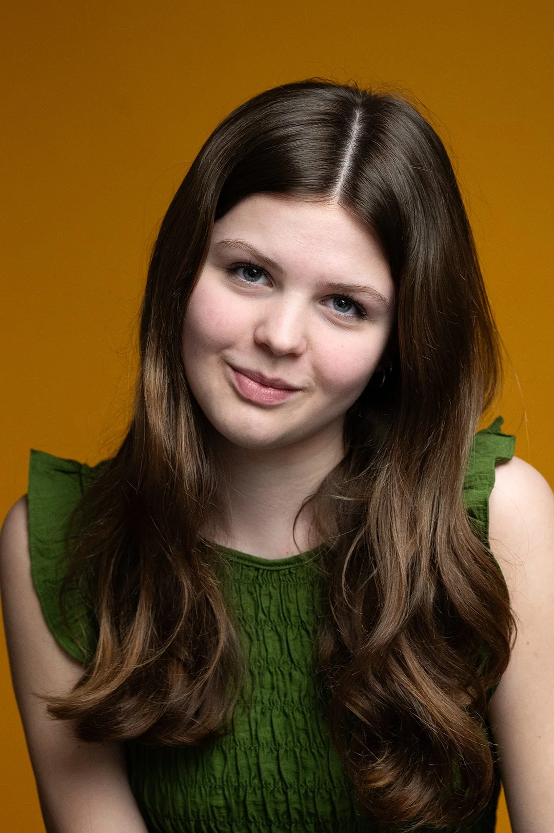 A young woman with long wavy brown hair and blue eyes, wearing a sleeveless green top, smiling gently against a solid yellow background.