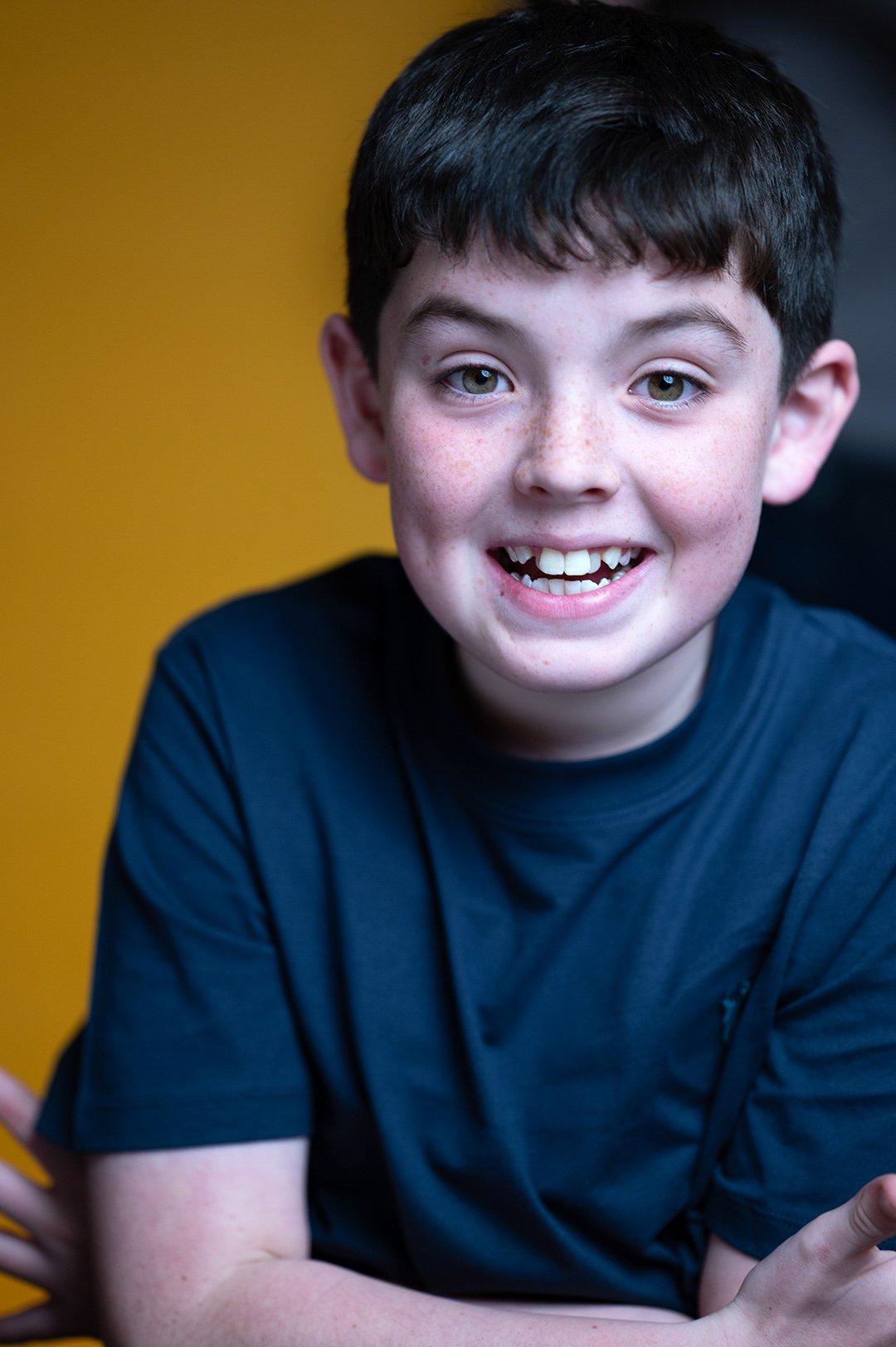 A young boy with dark hair and light skin with freckles, smiling with missing teeth, wearing a dark T-shirt, against a yellow background.