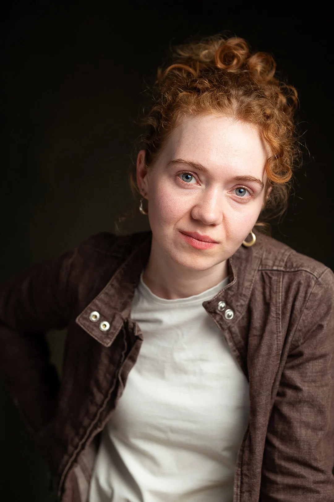 Portrait of a young woman with curly red hair and blue eyes, wearing a brown jacket and a white shirt, against a dark background.