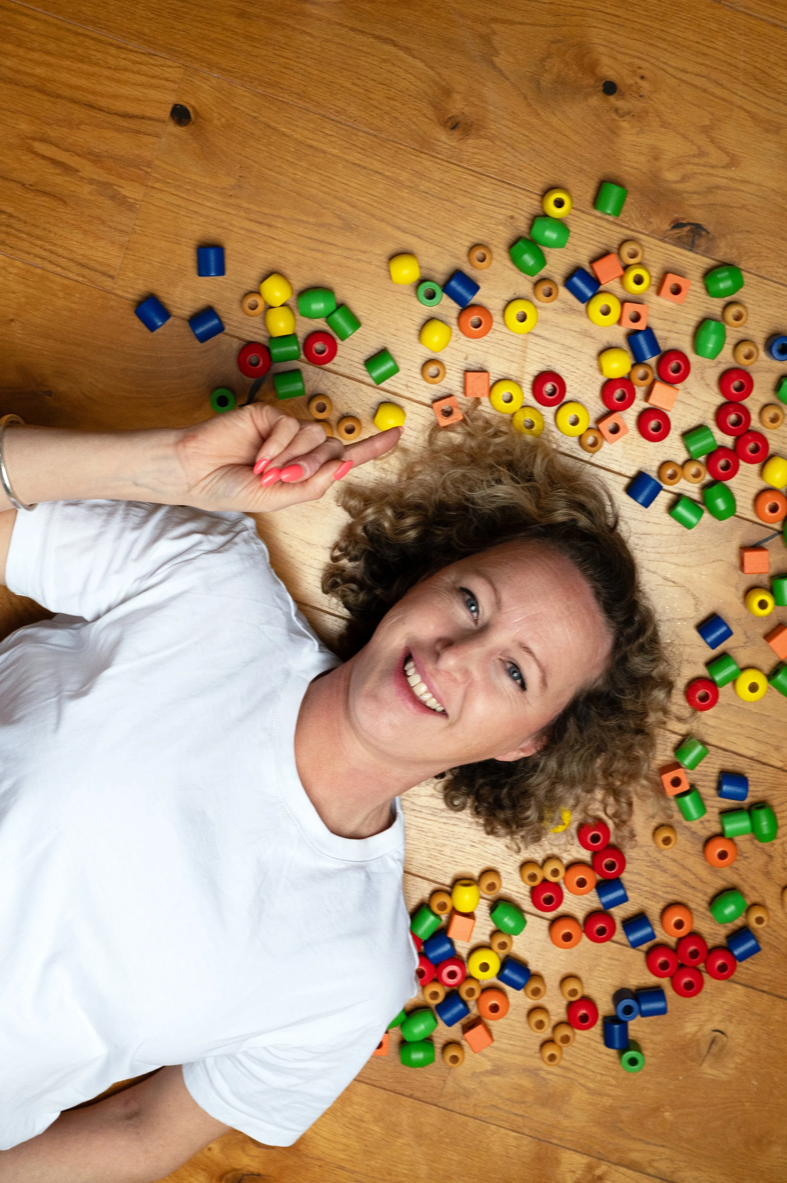 A woman lying on a wooden floor surrounded by colorful plastic beads, smiling at the camera.