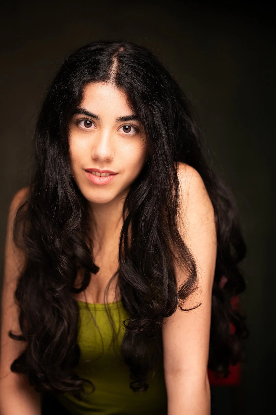 Young woman with long, black wavy hair and olive skin wearing a green top against a dark background, smiling slightly. 