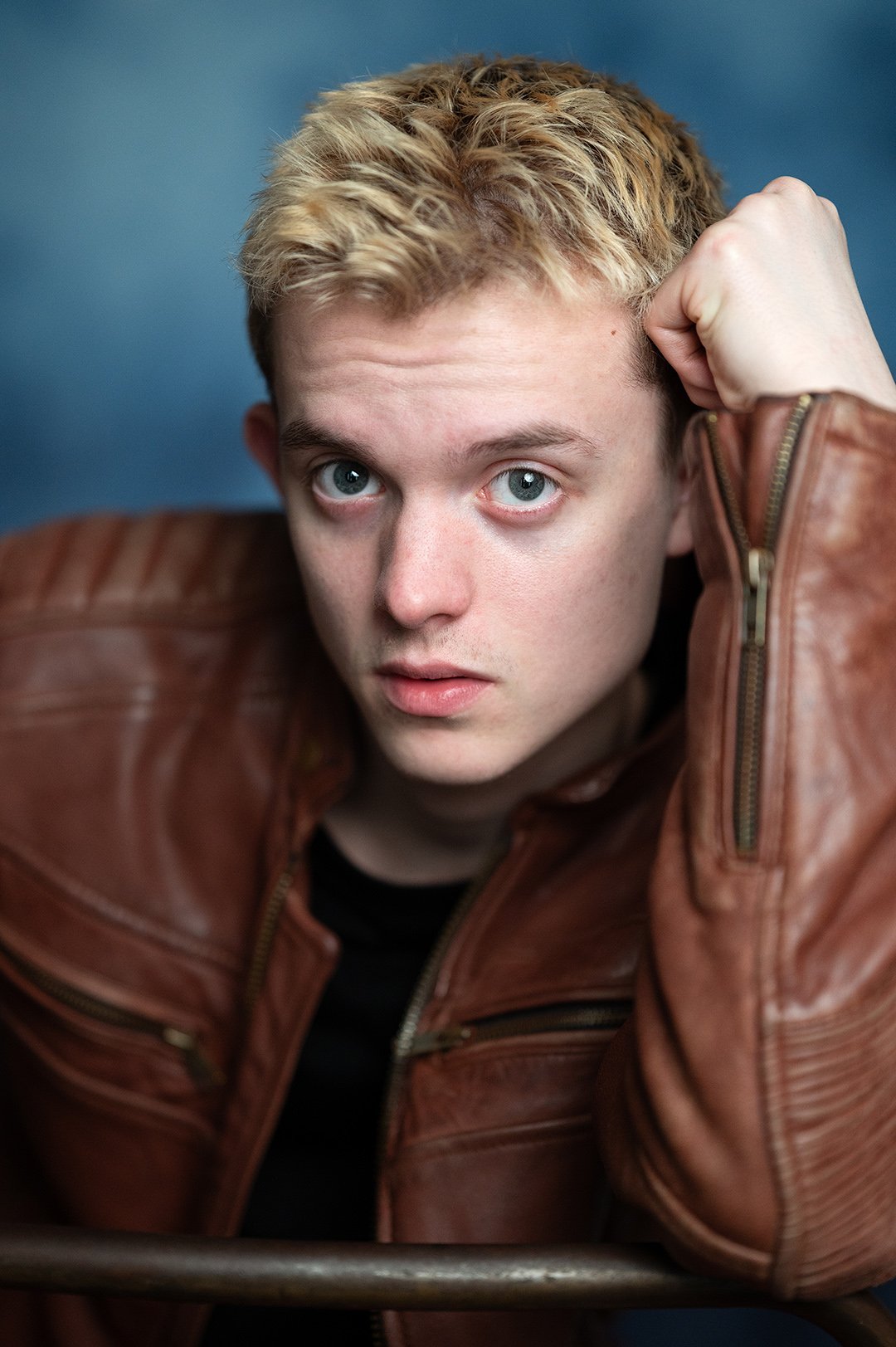 A young man with short blond hair and blue eyes wearing a brown leather jacket, posing with his hand resting on his head against a blue background.