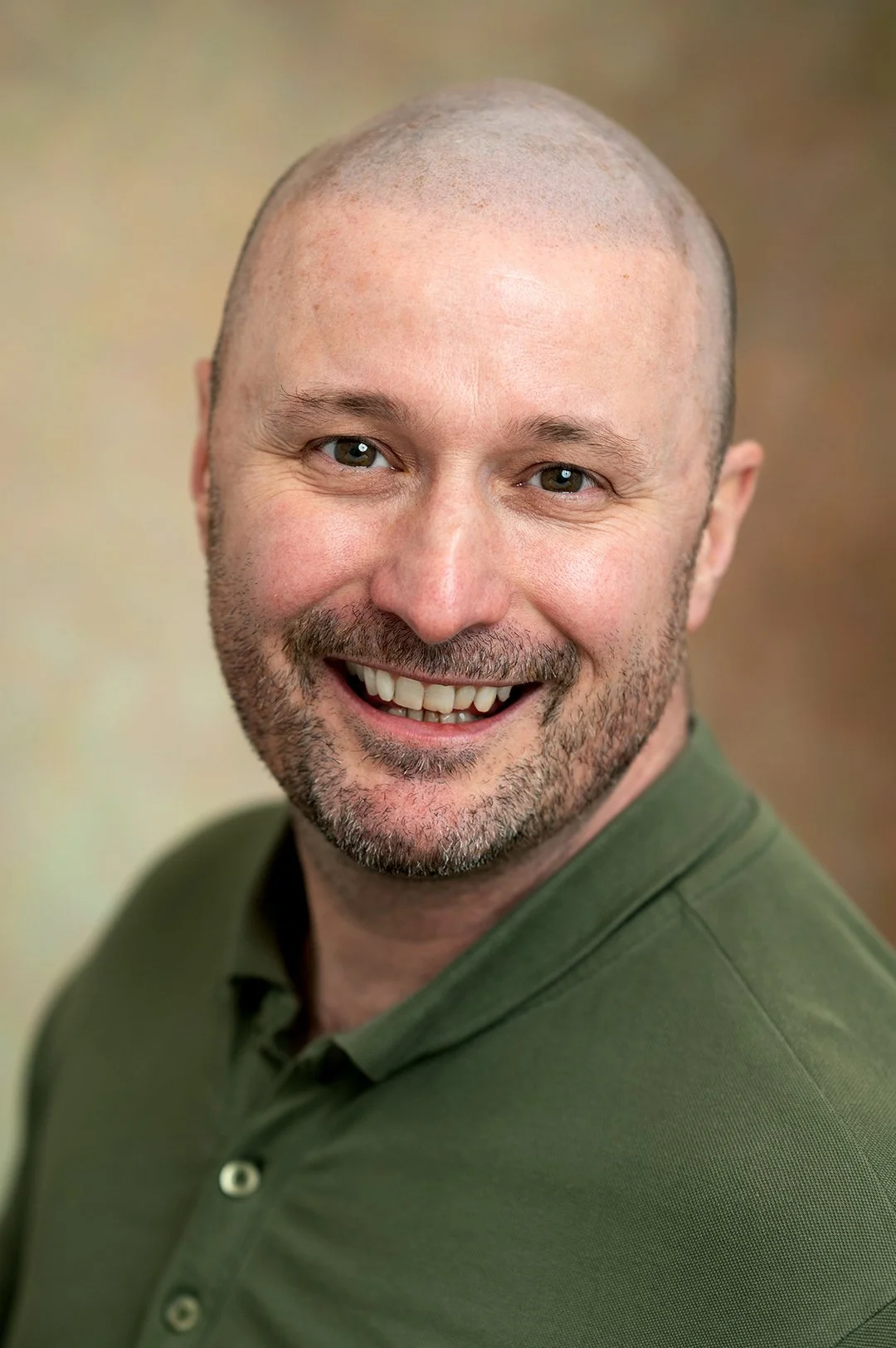 A male with a bald head photographed wearing a green top with a collar against a brown background. He is smiling and looking straight at the camera. 