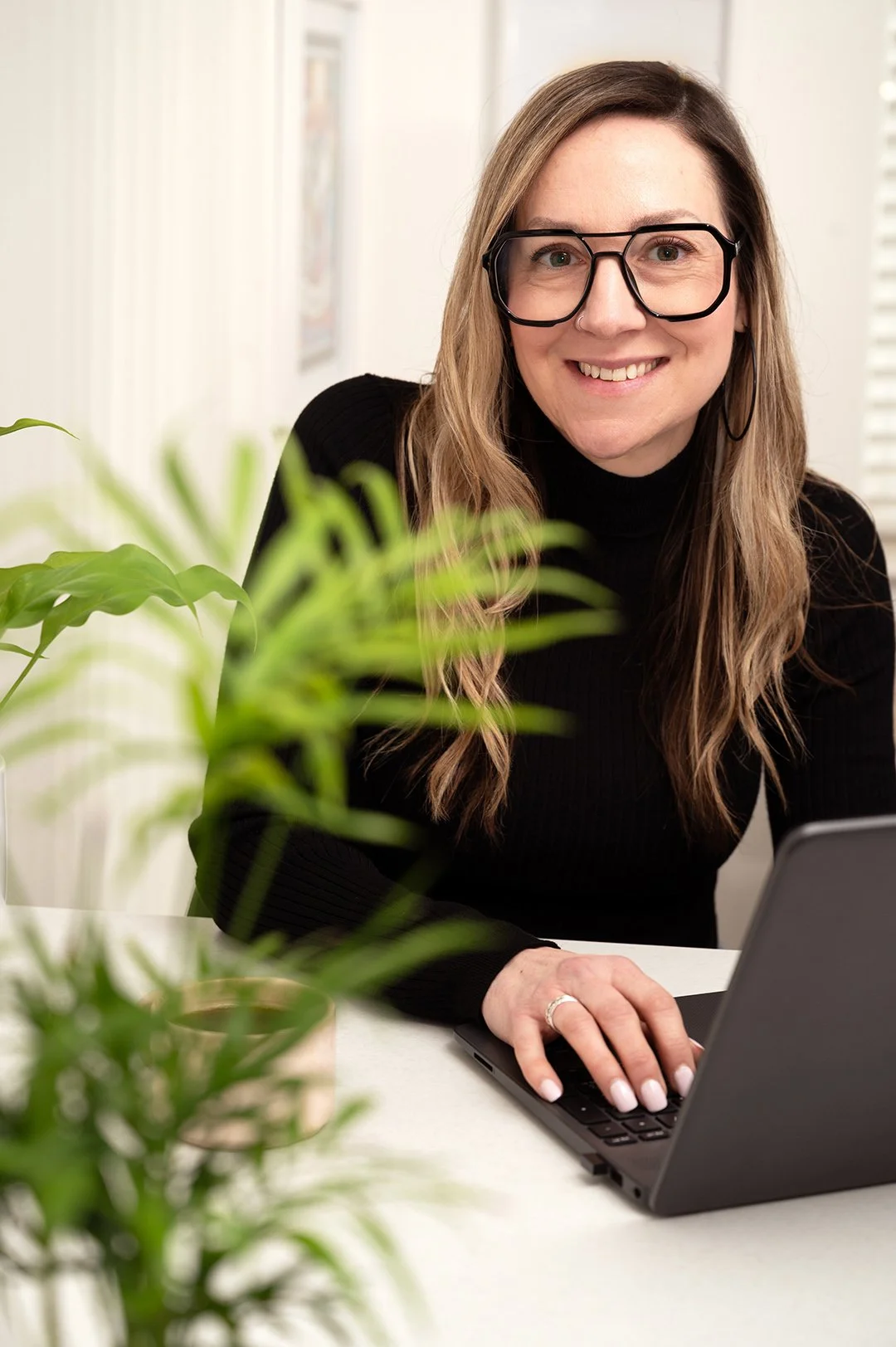 A woman with long wavy hair, wearing glasses and a black sweater, smiling while working on a laptop at a white desk. Green houseplants and a blurred background are visible.