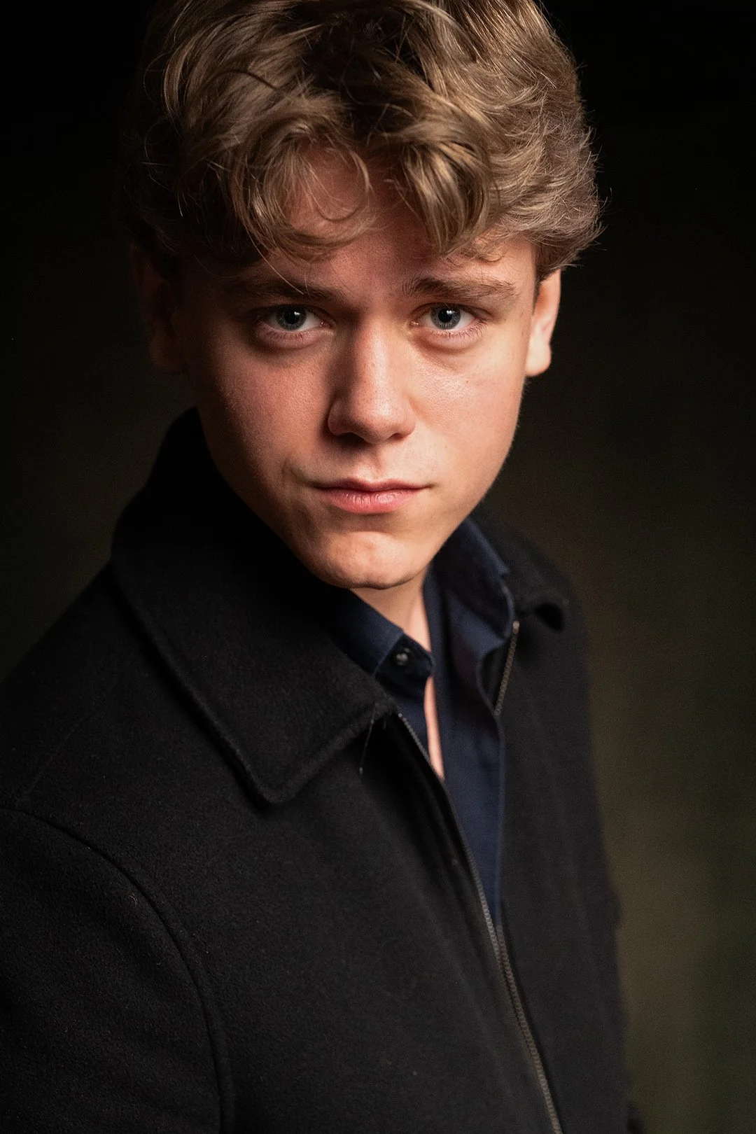 A young man with a dark jacket and blue shirt posing with his shoulder and head facing straight at the camera. The background is dark. His expression is determined. 