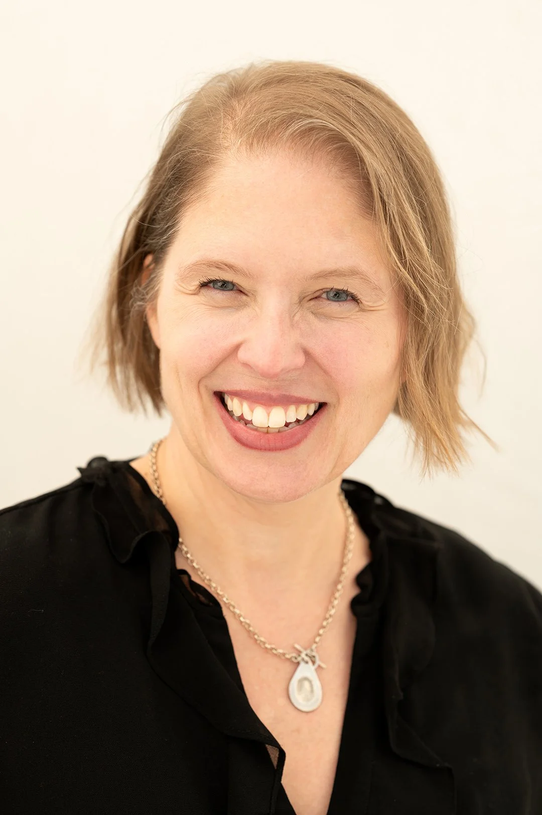 A female entrepreneur smiling straighth at the camera wearing a black blouse, with a necklace round her neck. the backdrop is white. 
