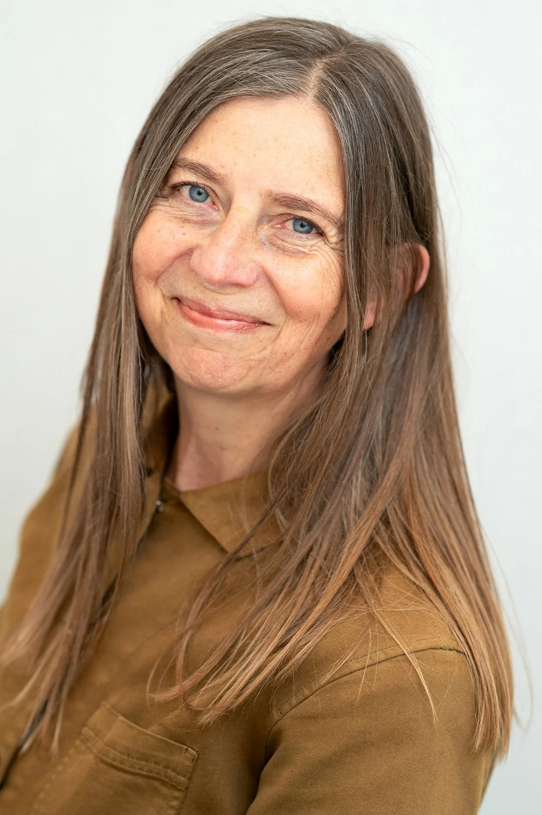 A woman with long straight brown hair wearing a brown jacket smiling straight at the camera. the backdrop is white. 
