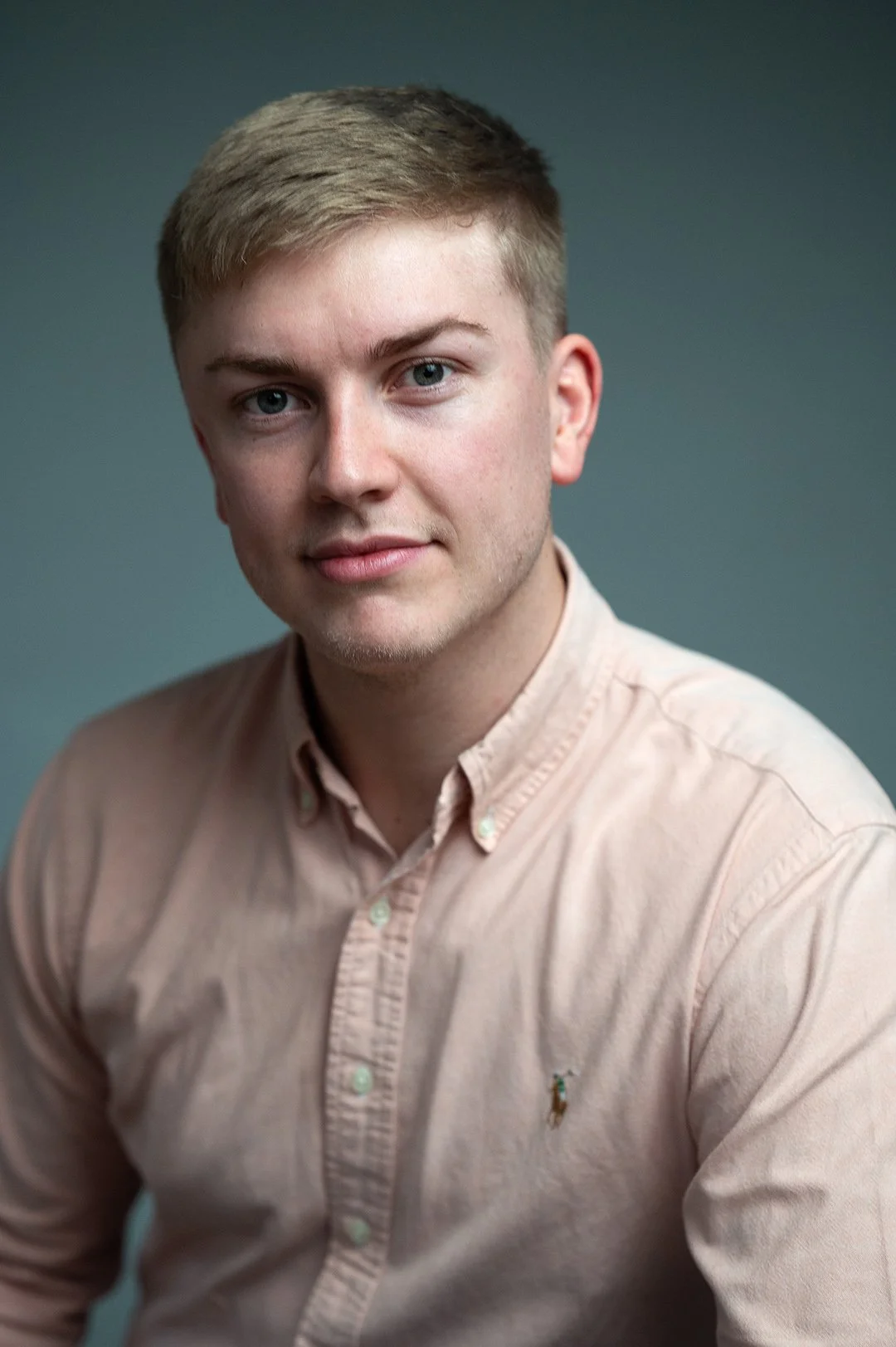 A young man with blonde hair wearing a beige button-up shirt against a plain gray background.