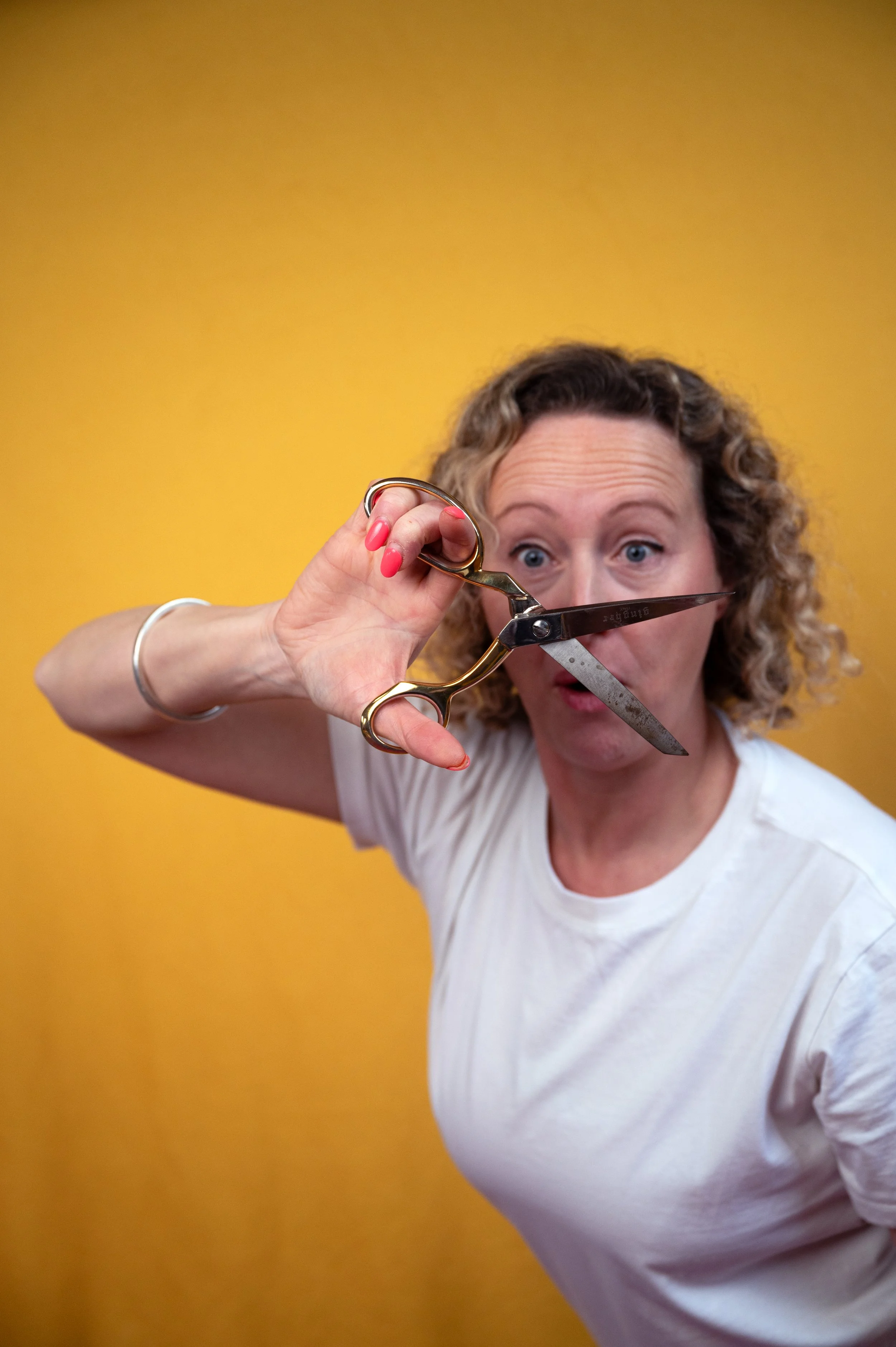 A woman with curly hair and a white shirt holds a pair of scissors in front of her face, with a surprised expression, against a yellow background.