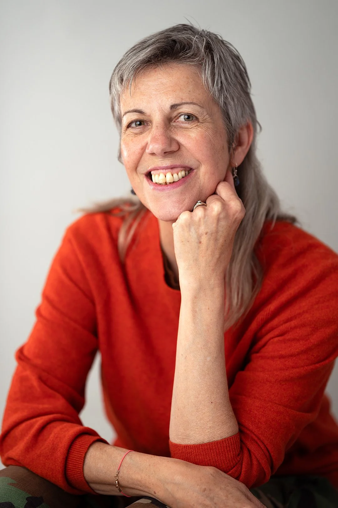 Woman wearing orange jumper smiling at the camera with grey long hair. Photographed against a light grey backdrop. 