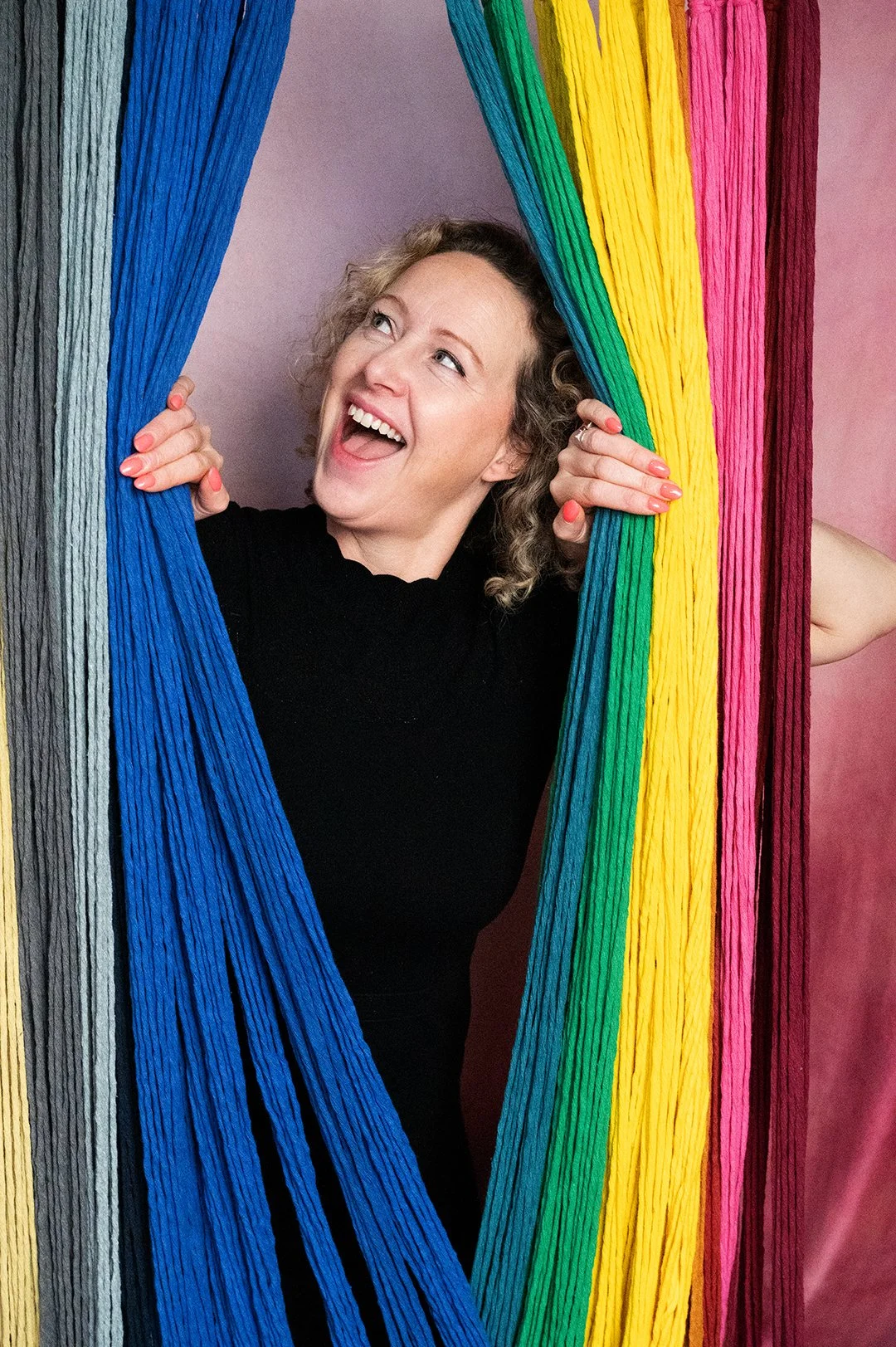 A woman peeks through a colorful string curtain with rainbow colors, smiling and looking joyful.