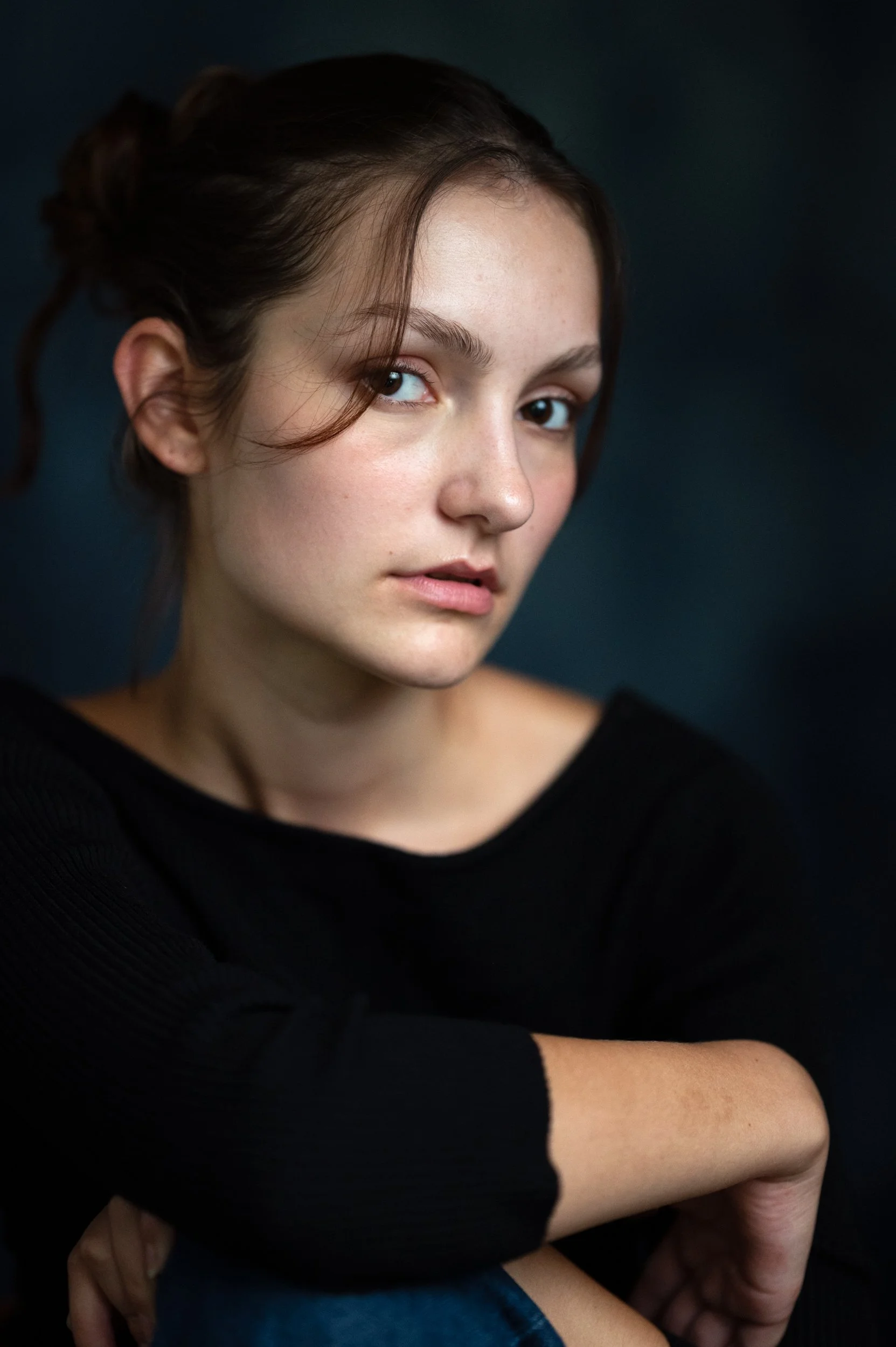 Close-up of a young woman with brown hair styled in a bun, wearing a black ribbed sweater, looking over her shoulder against a dark background.