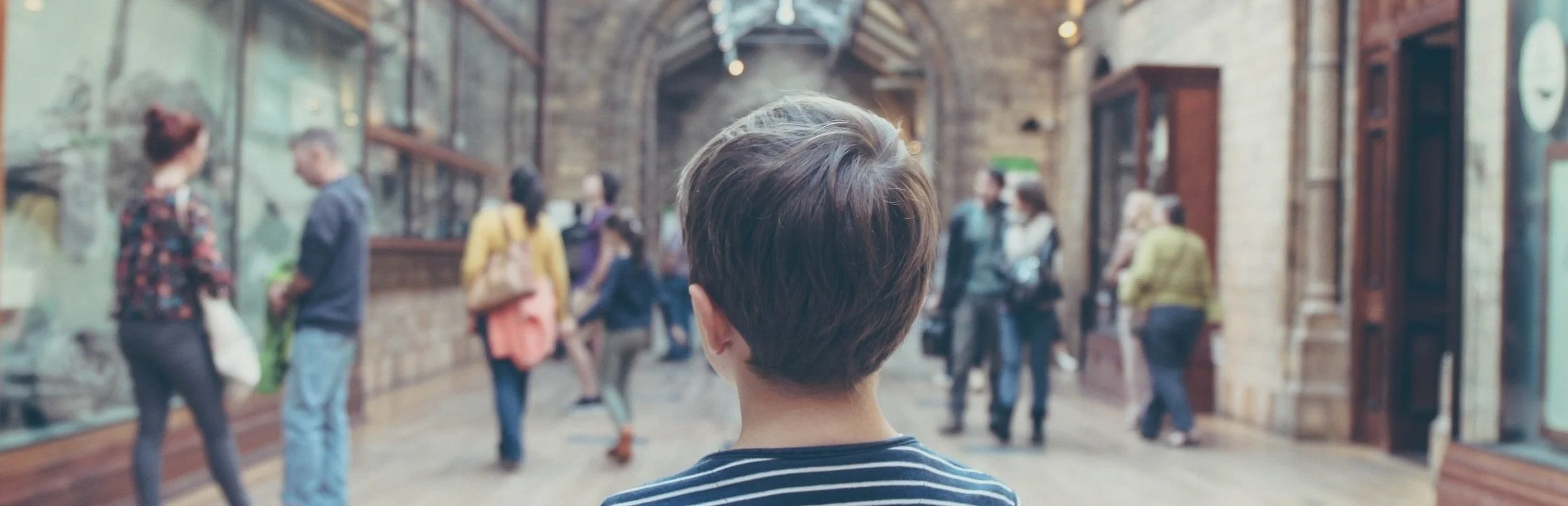 A view of a museum corridor looking from behind a young boy into the space where people are walking, talking, and looking at exhibits. Michal Parzuchowski