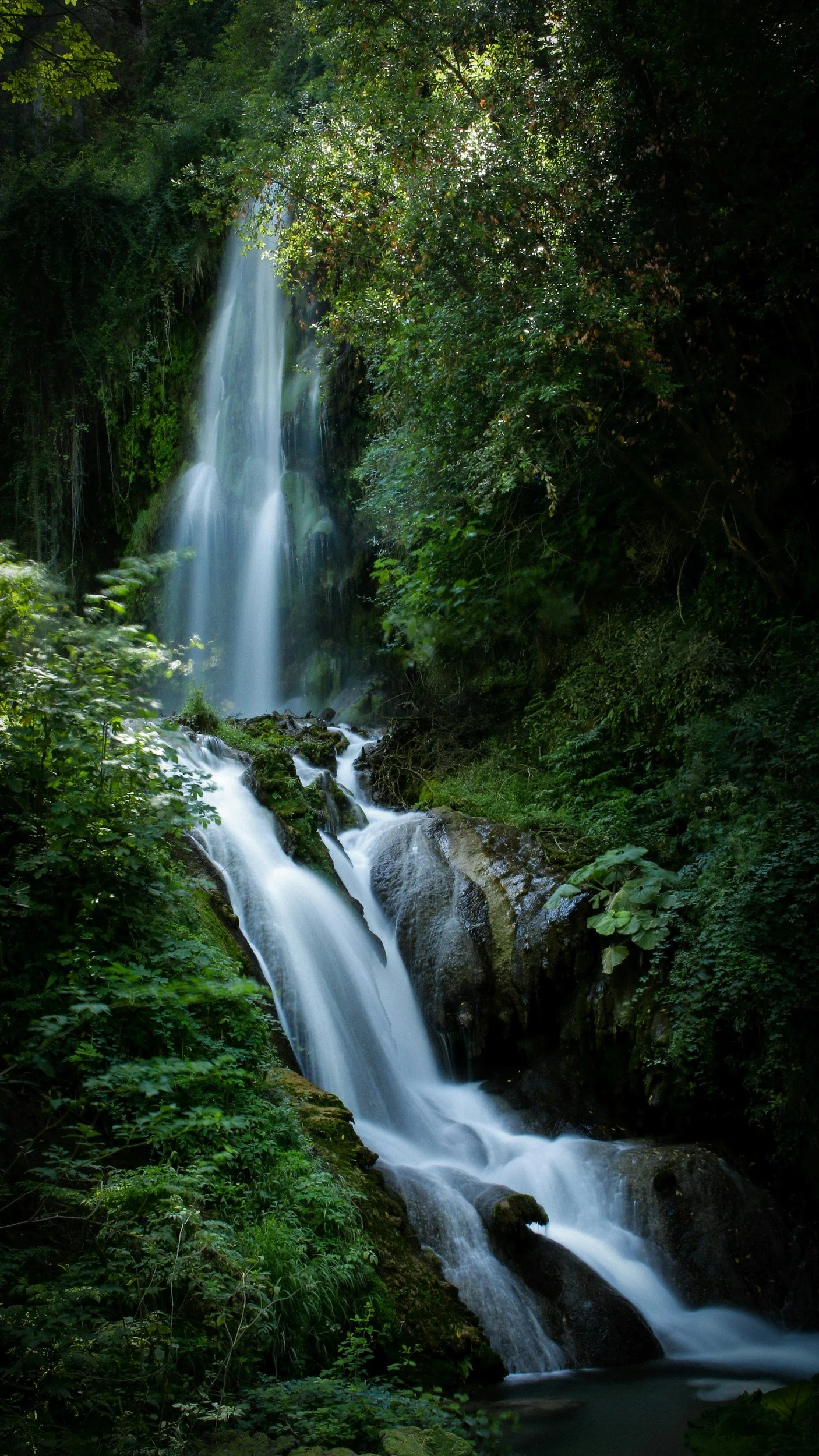 A waterfall flowing downhill through lush green trees and vegetation.