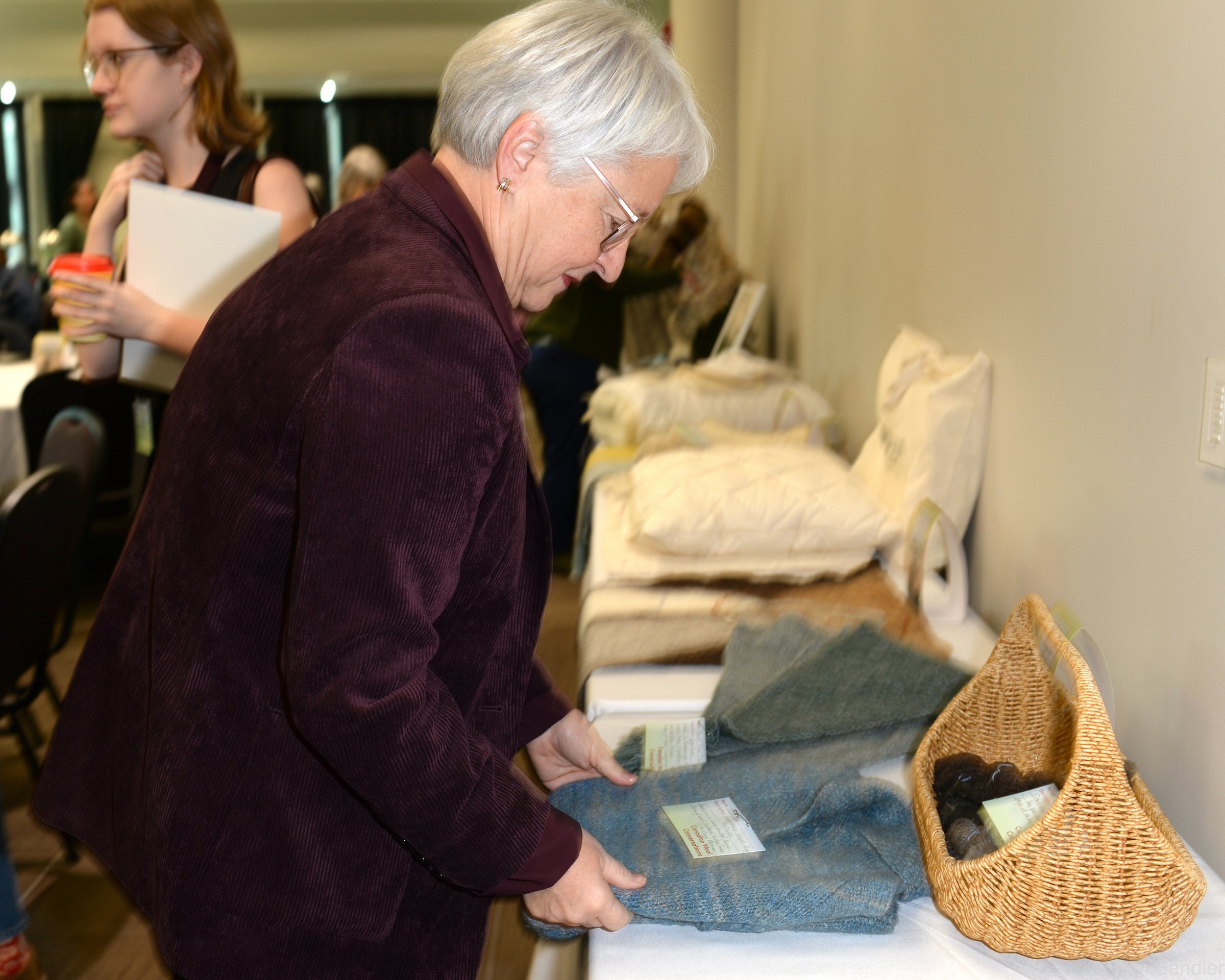 Une femme âgée aux cheveux blancs, portant des lunettes et vêtue d'une veste marron, examine des écharpes ou des châles en laine exposés sur une table dans un salon ou un marché artisanal, avec à côté d'elle un panier en osier contenant de la laine ou du fil.
