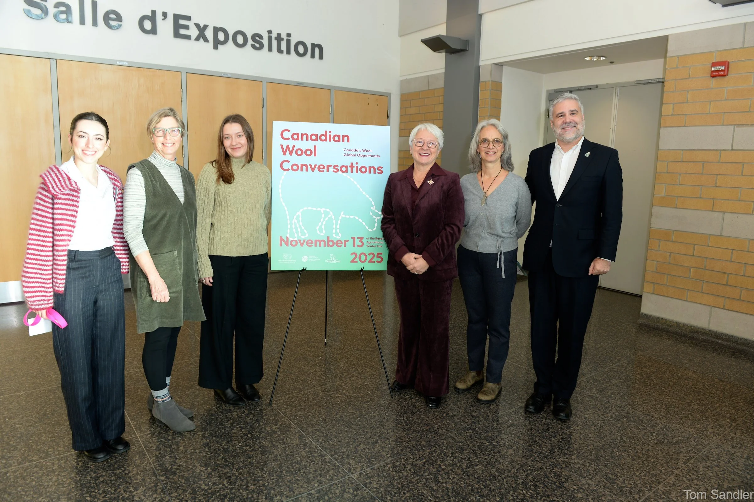 Sept personnes debout en file devant un panneau sur lequel on peut lire « Canadian Wool Conversations » (Conversations sur la laine canadienne) avec la date du 13 novembre 2025.