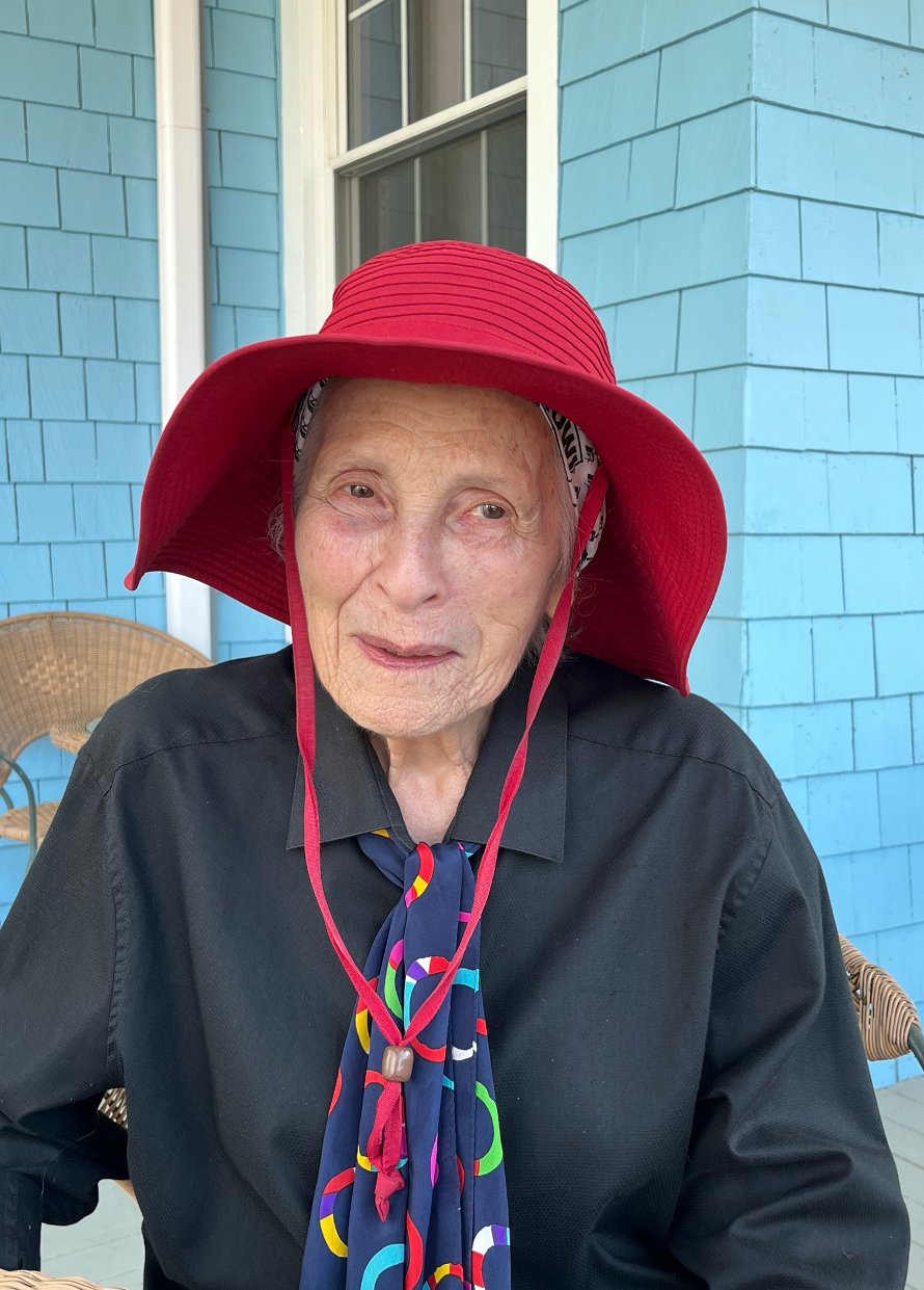 The author in a vibrant red hat, dark blouse with a colorful tie, sitting in front of a house with light blue shingles.
