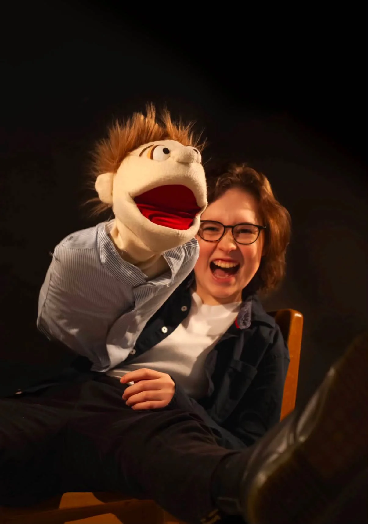 A woman with glasses and short brown hair laughing while sitting on a chair, with a puppet that has a large mouth, brown spiky hair, and a striped shirt, positioned next to her against a dark background.