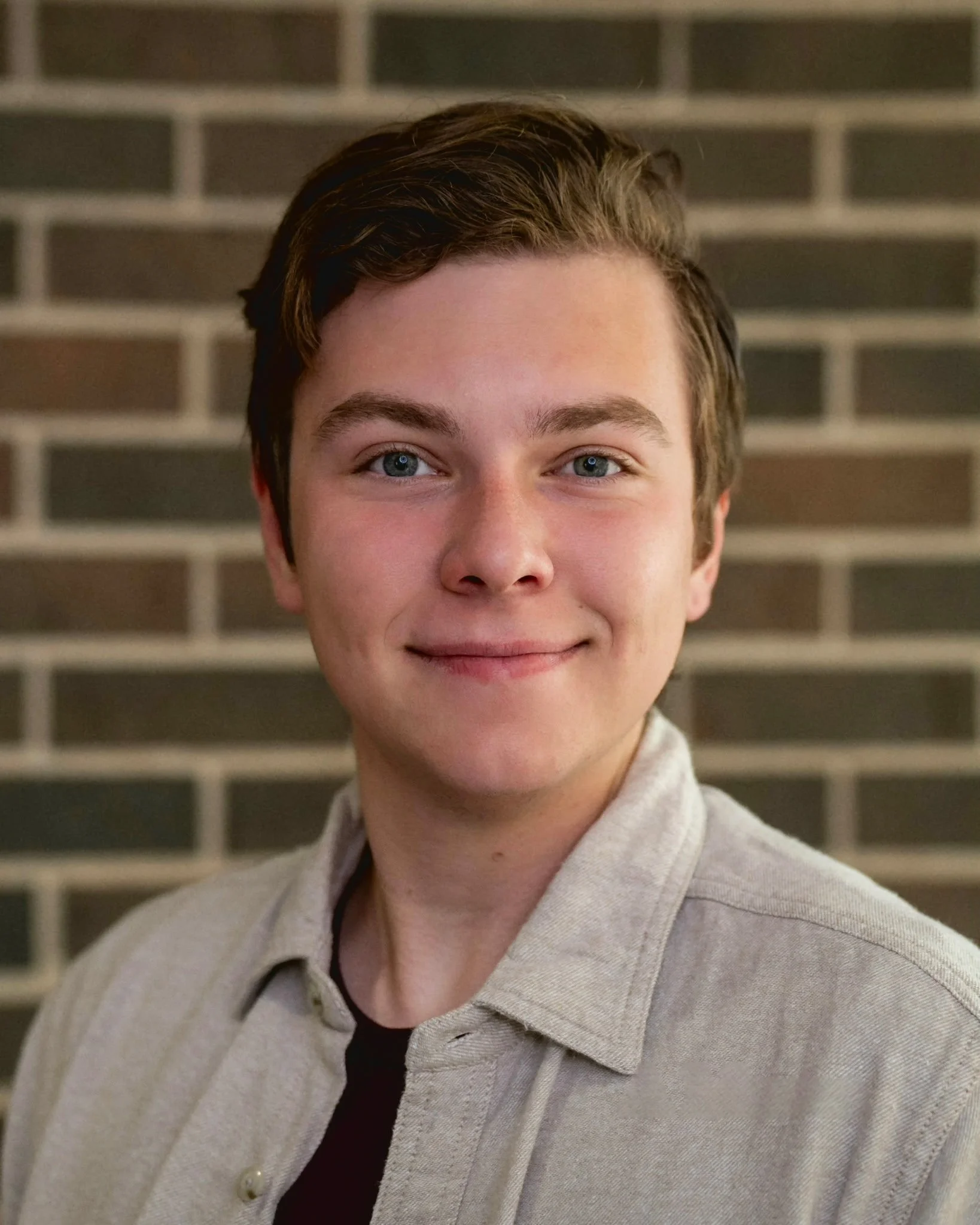 A young person with short brown hair, blue eyes, and fair skin, wearing a beige jacket and a black shirt, standing in front of a brick wall.