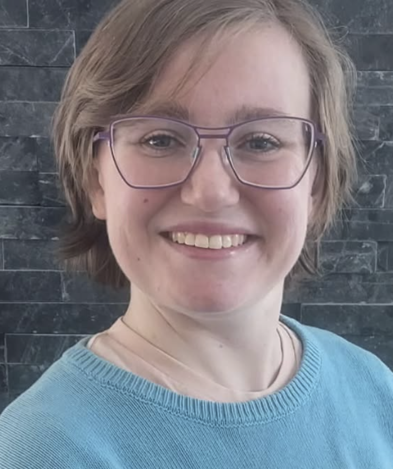 A woman with short light brown hair, wearing glasses with purple frames, smiling, dressed in a light blue top, standing against a dark stone wall background.