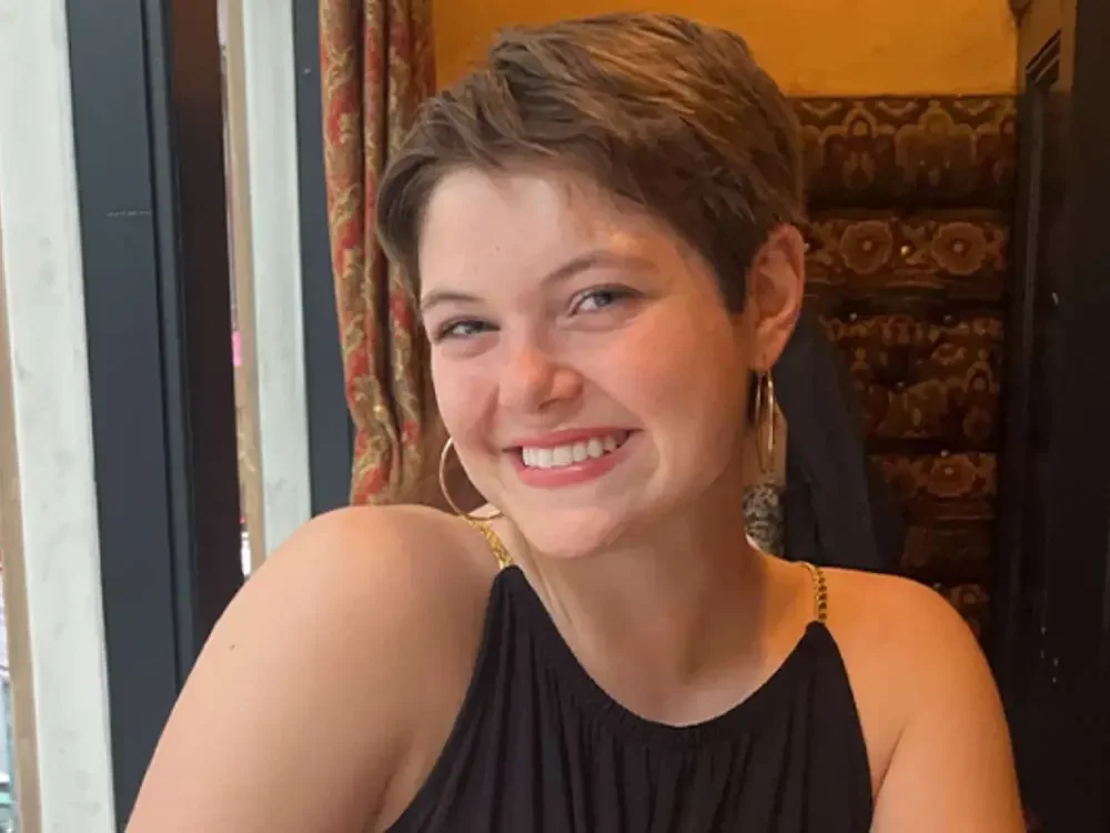 A young woman with short brown hair smiling and wearing a black sleeveless top, gold earrings, and a small gold chain, sitting in a restaurant with patterned curtains and a wooden wall behind her.