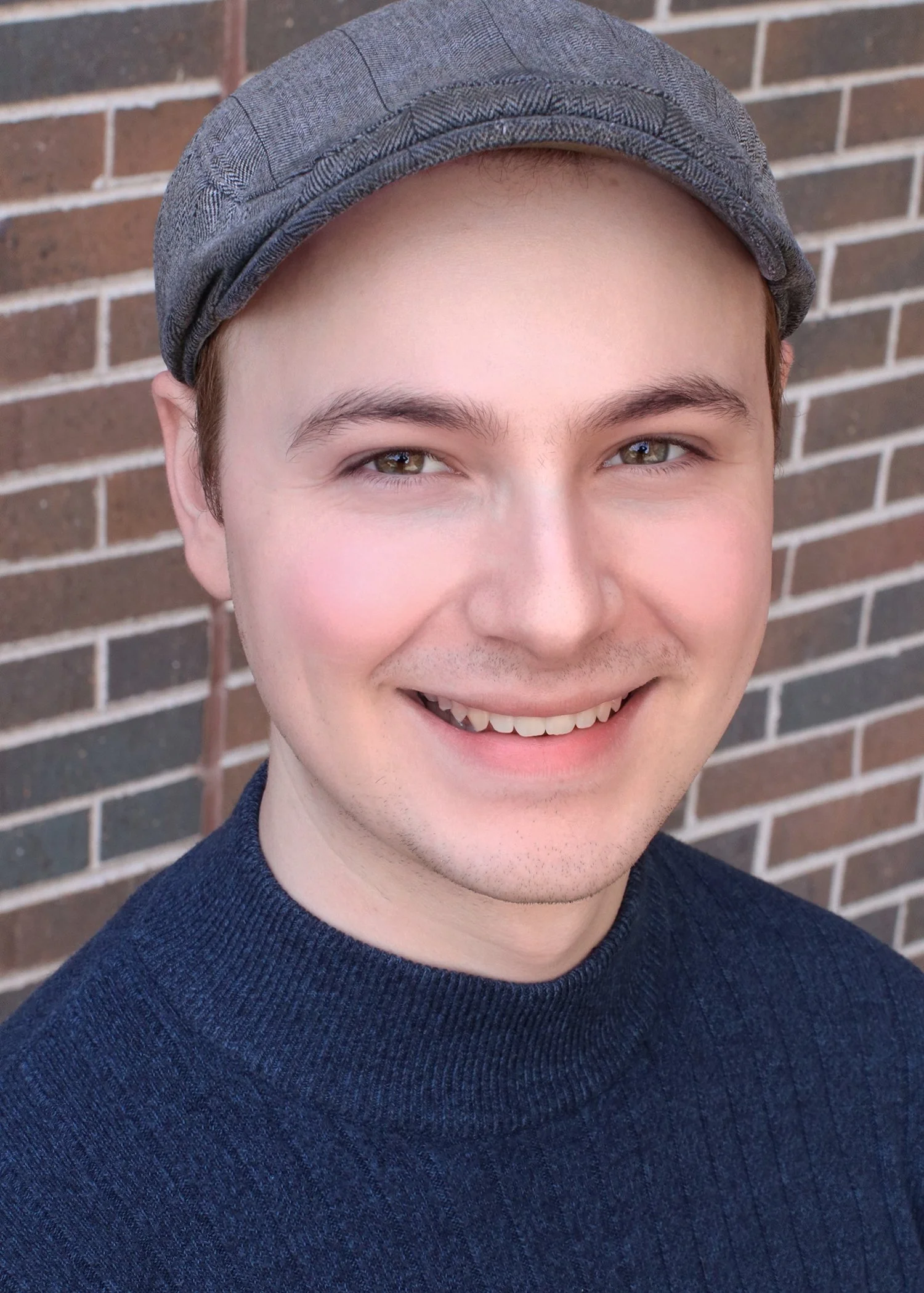 Close-up of a young man smiling, wearing a gray flat cap and a navy blue sweater, standing in front of a brick wall.