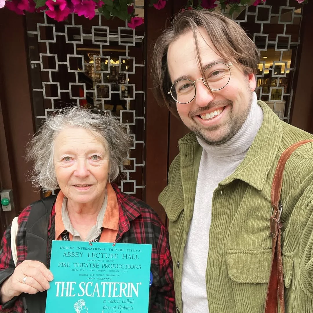 A woman with gray, curly hair holding a blue program for 'The Scatterin'' play, standing beside a smiling man with glasses and brown hair, outside a building with decorative metalwork and pink flowers above.